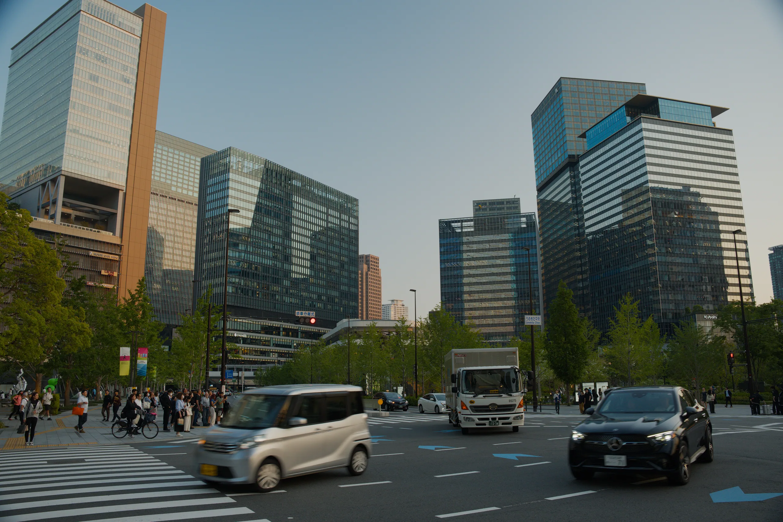 Busy urban intersection with tall office buildings and pedestrians crossing at a marked crosswalk.