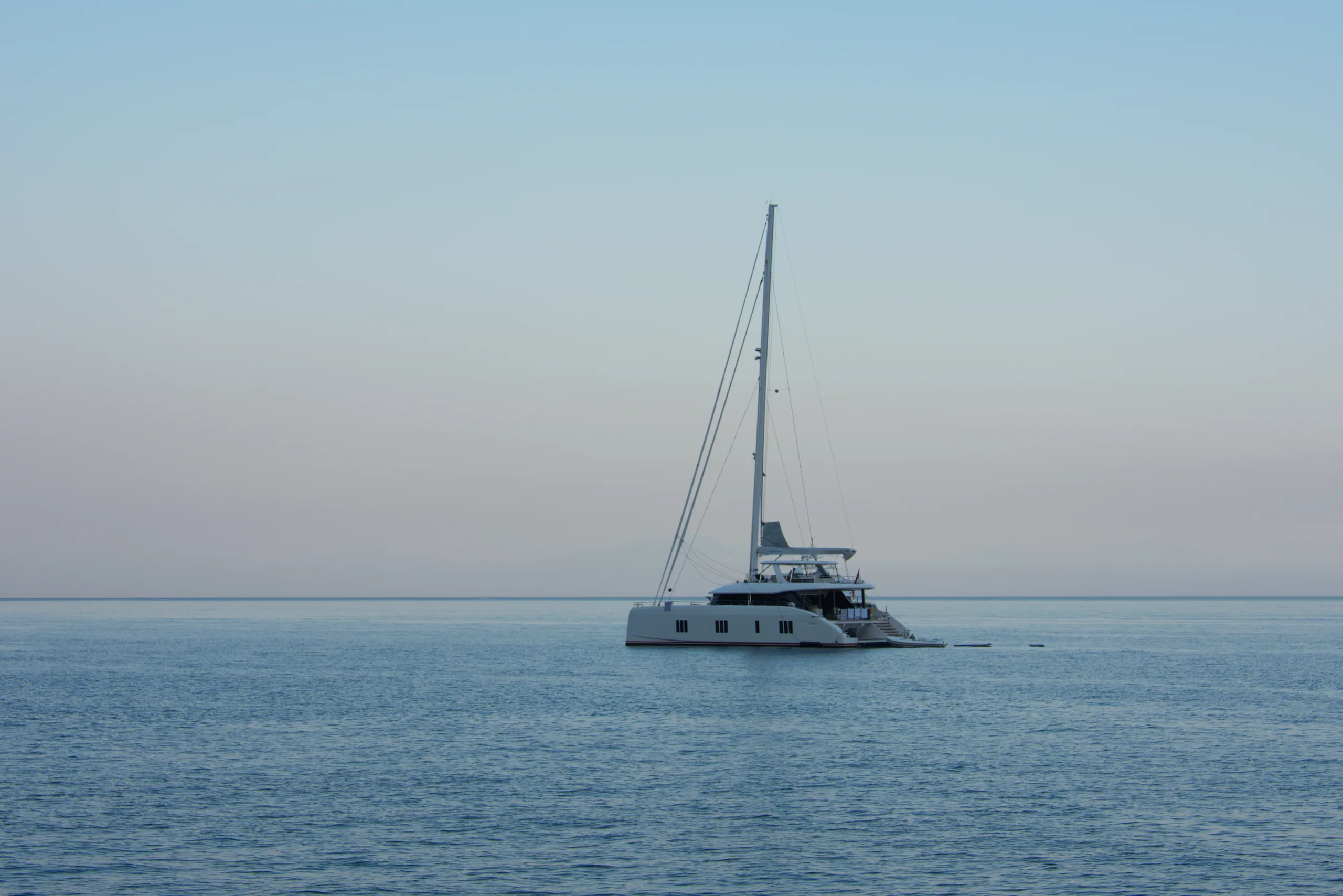 A white catamaran under bare mast on a flat calm sea, the horizon hazing into the sky.