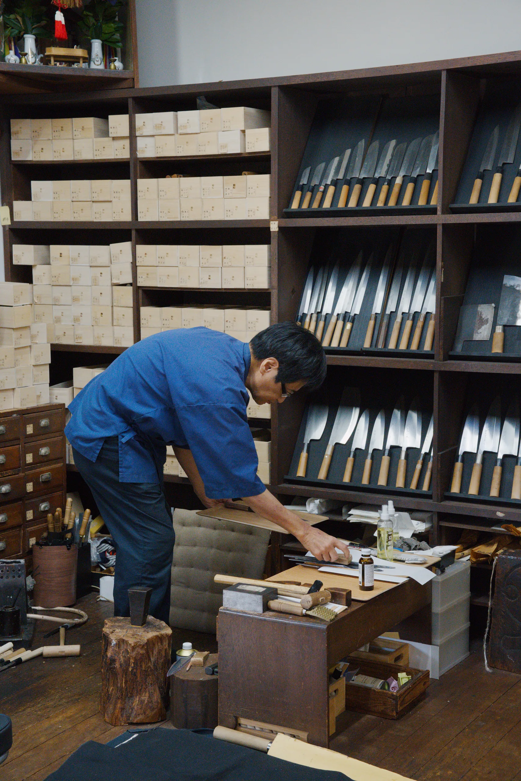 Person in blue work clothes standing at a wooden station working with hand tools and wood blocks.