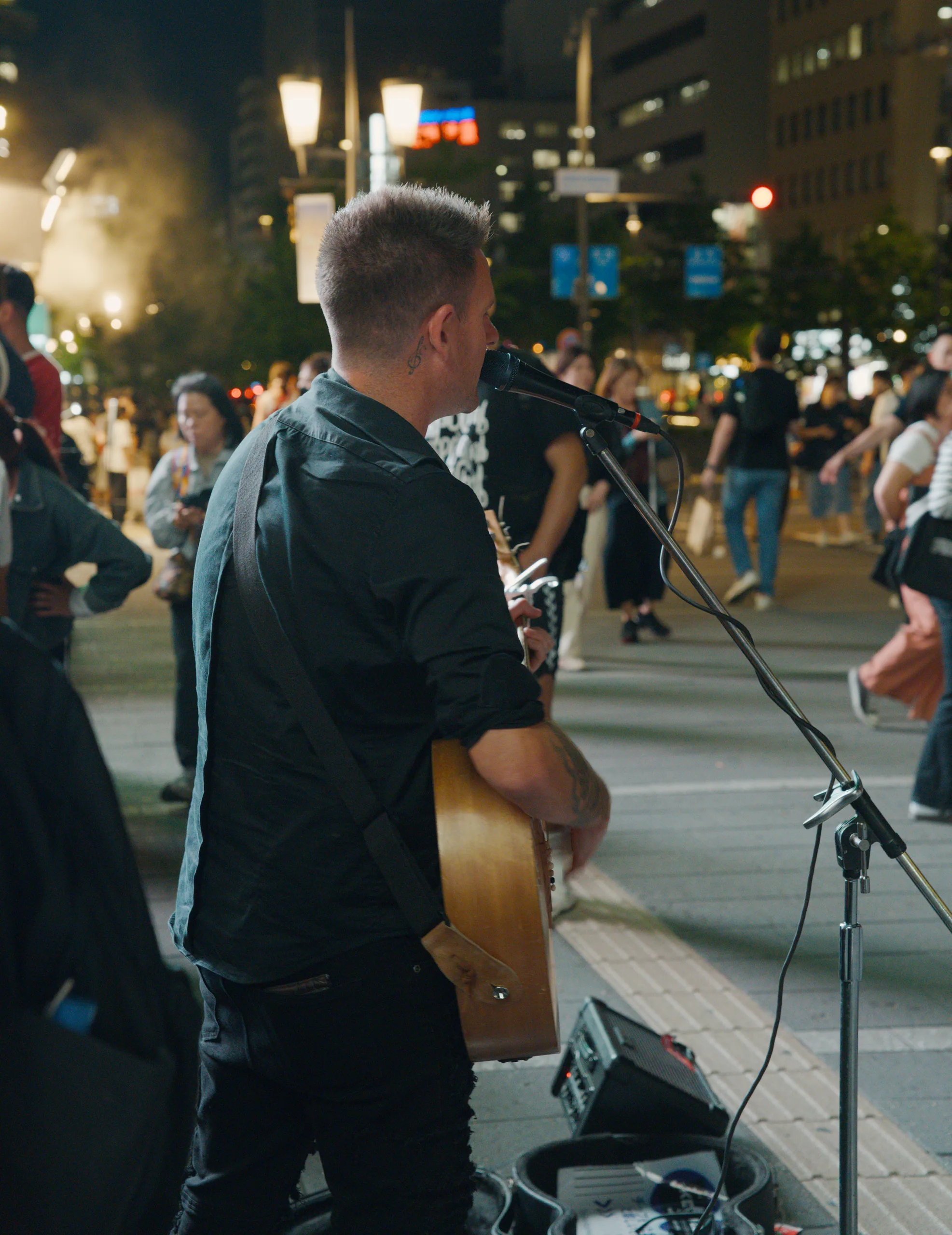 Street musician with guitar performing at night on an urban street with city lights beyond.