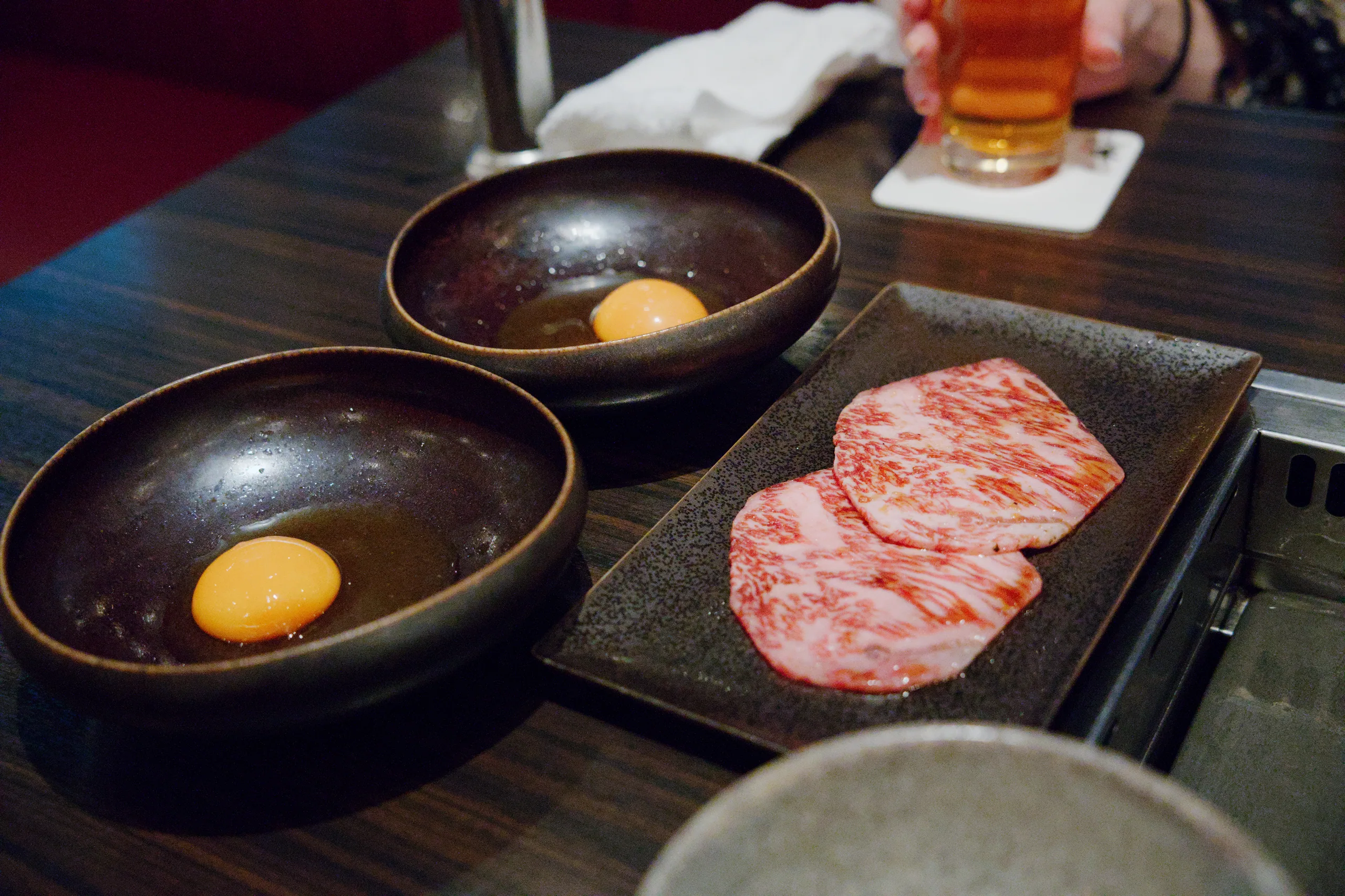 Multiple dark bowls hold soft egg yolks and thin slices of cured meat arranged at a restaurant counter.