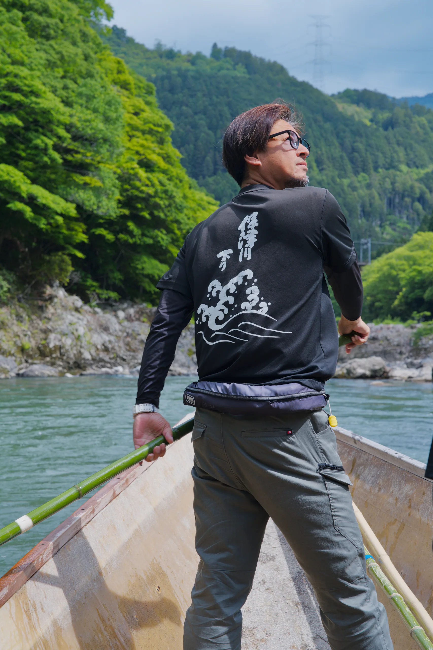 A person poles a boat across turquoise water beneath forested mountains.