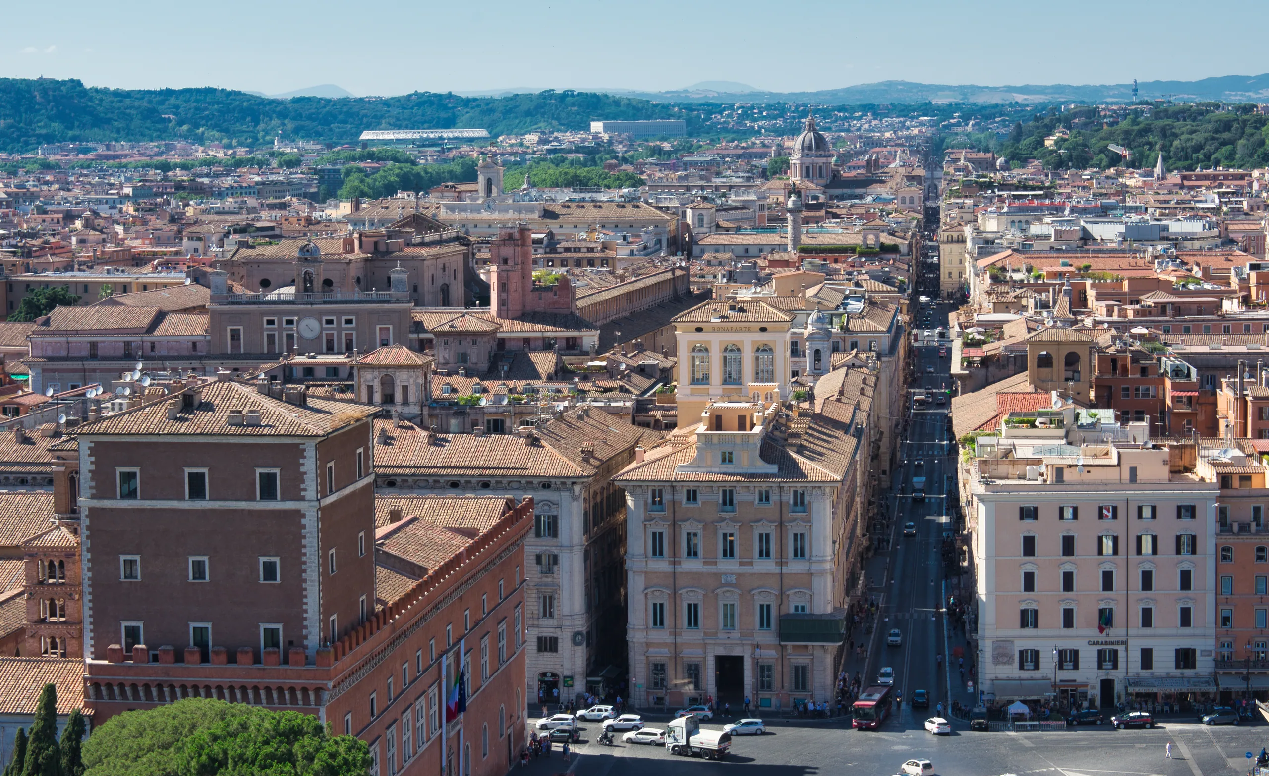 A high view down Via del Corso, straight between rooftops and palazzi, with the hills beyond the city on the horizon.
