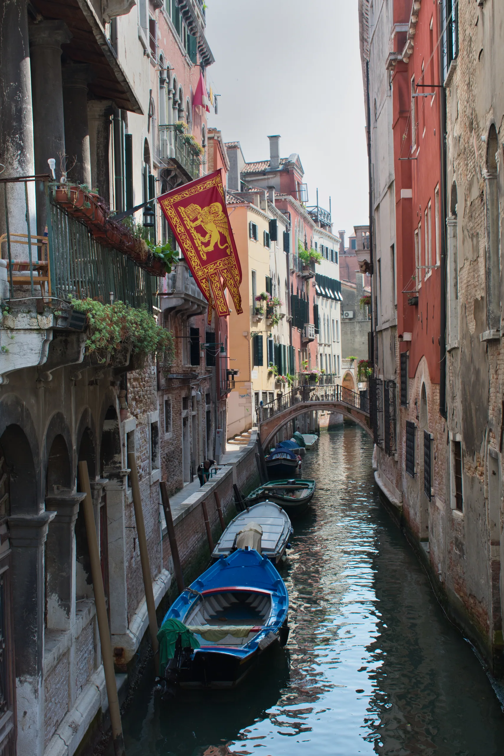 A Venetian canal with a gold and red banner hanging from a balcony, a blue wooden boat moored below.