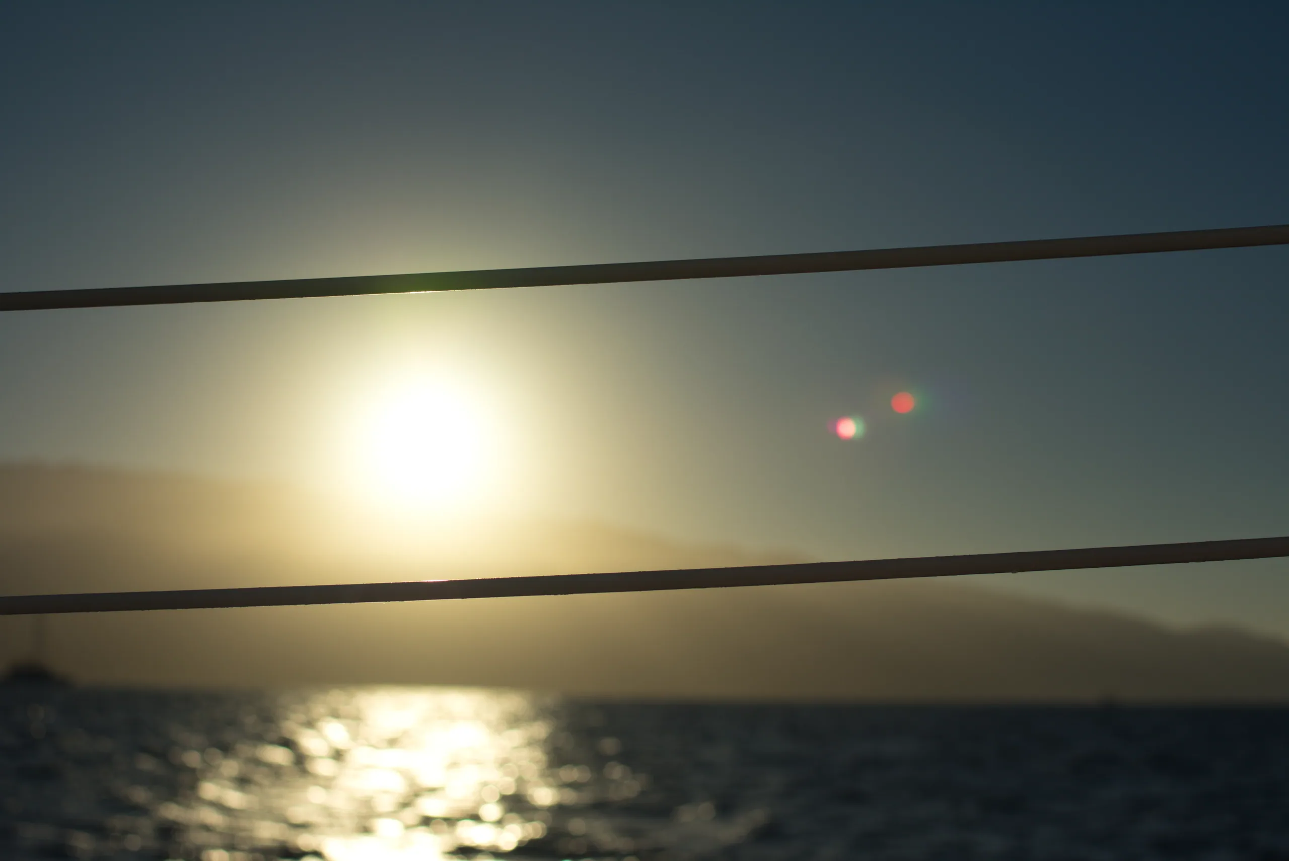 The sun low over the sea behind two power lines, its light laid in a bright path across the water.
