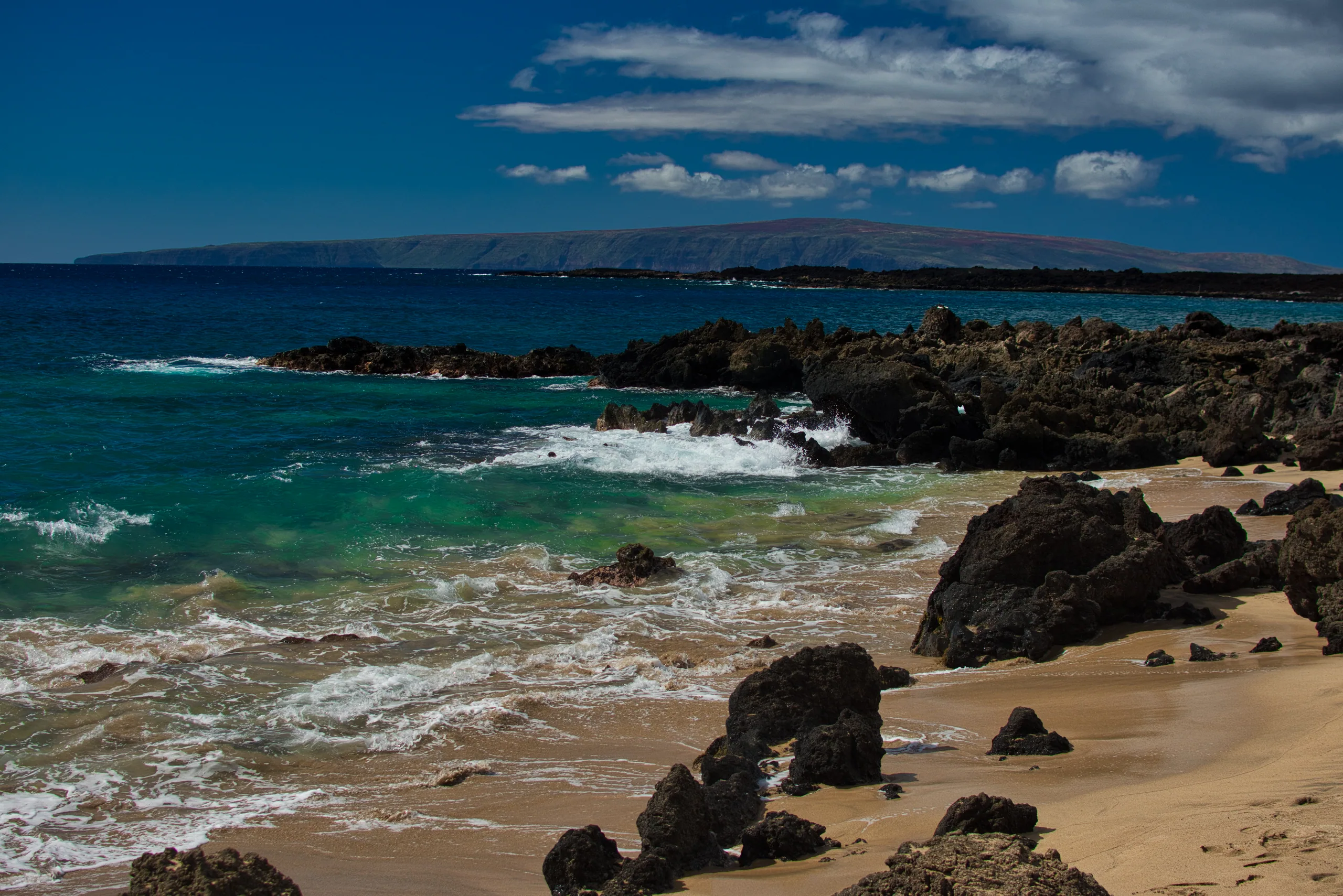 A crescent of pale sand between black lava outcrops, turquoise surf washing in and a long island profile on the horizon.