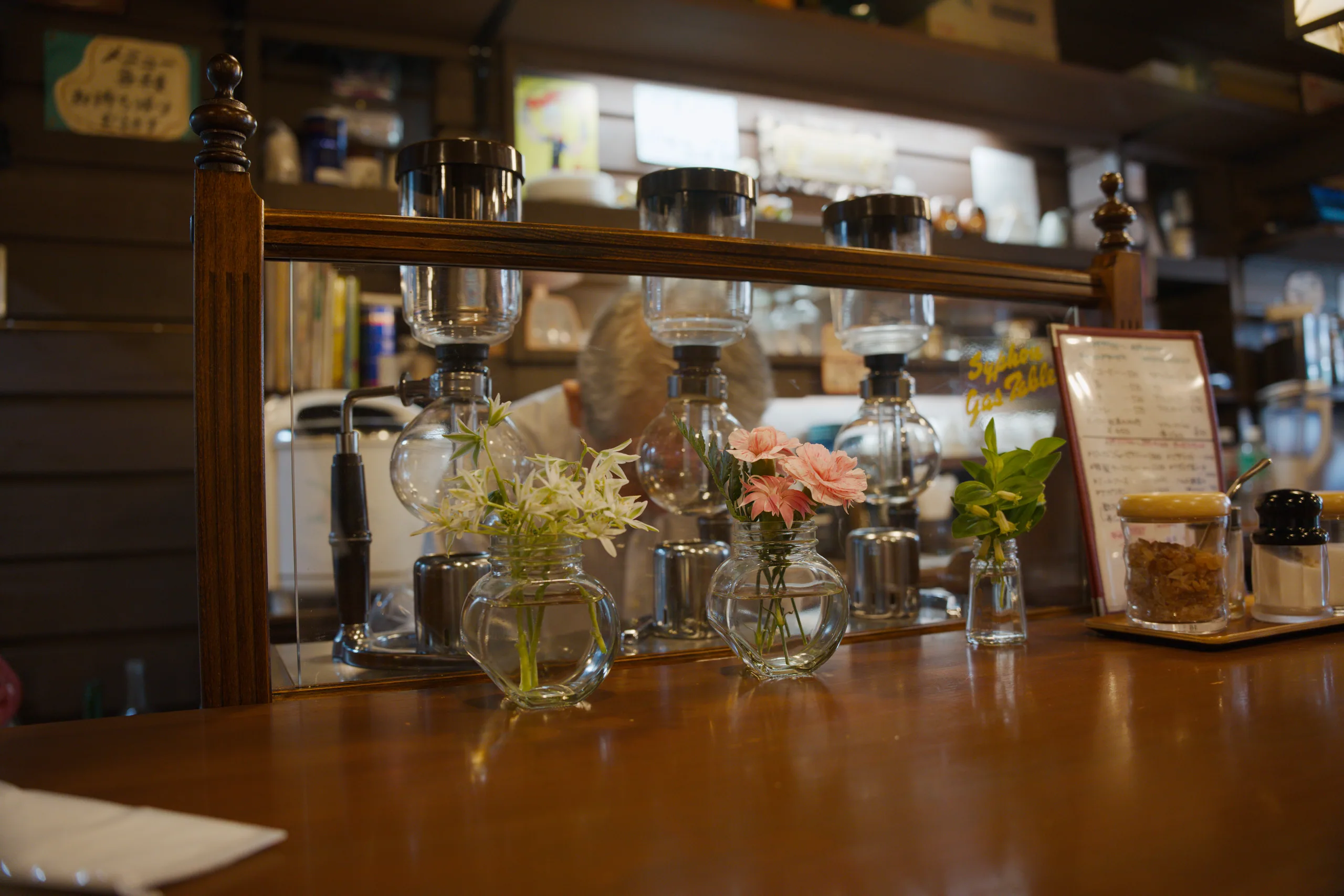Fresh flowers in glasses line a wooden bar counter with shelves of bottles behind.