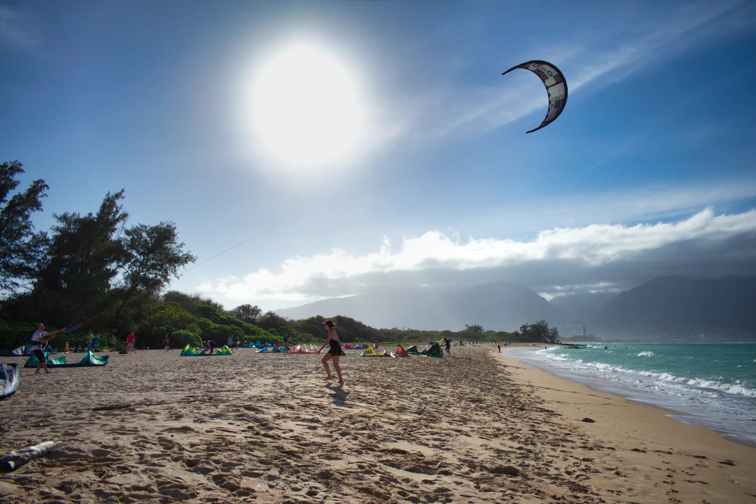 A kitesurfer's canopy high against a bright sun over a windswept beach, kites rigged along the sand below.