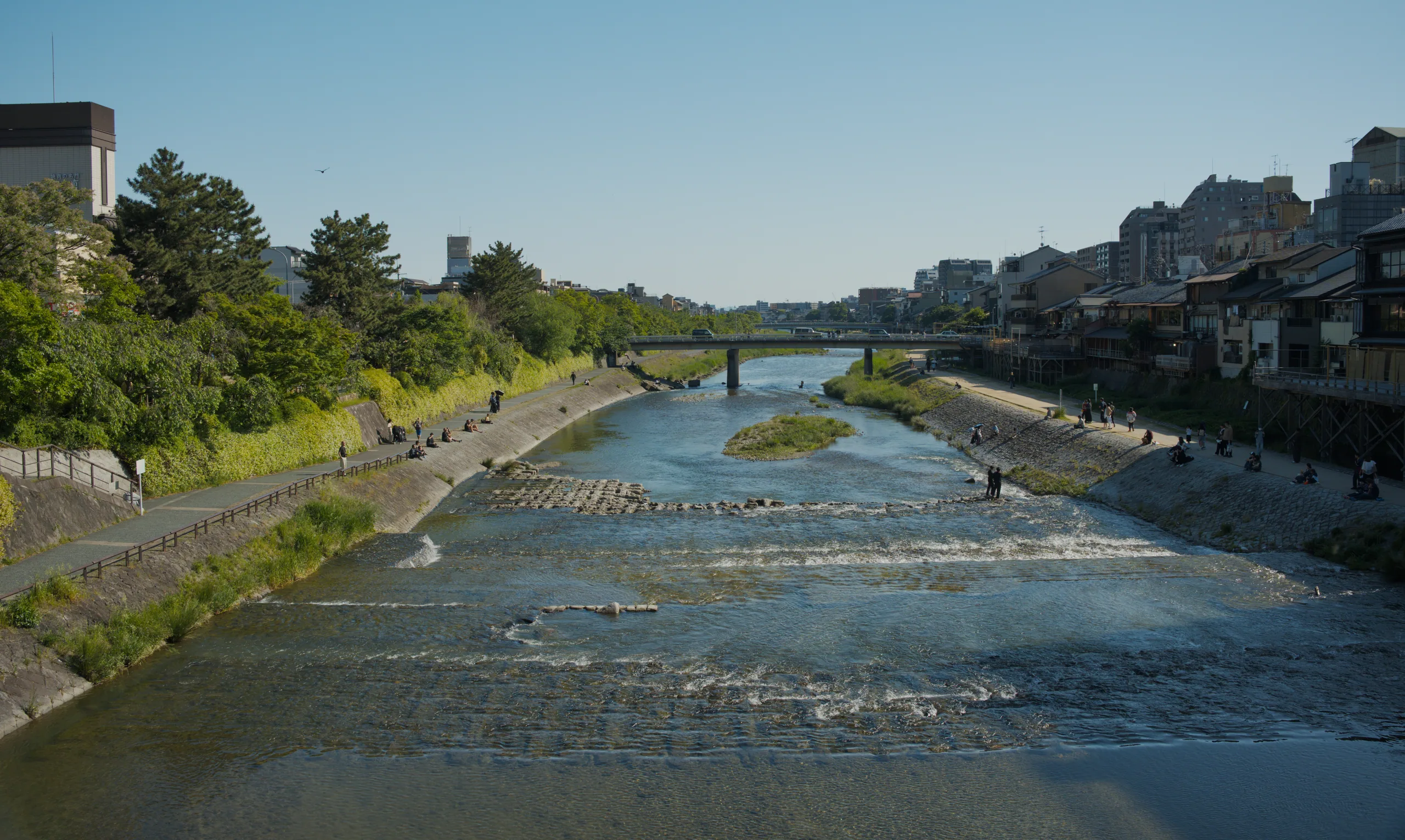 A calm river bordered by grassy banks and city buildings under clear sky.