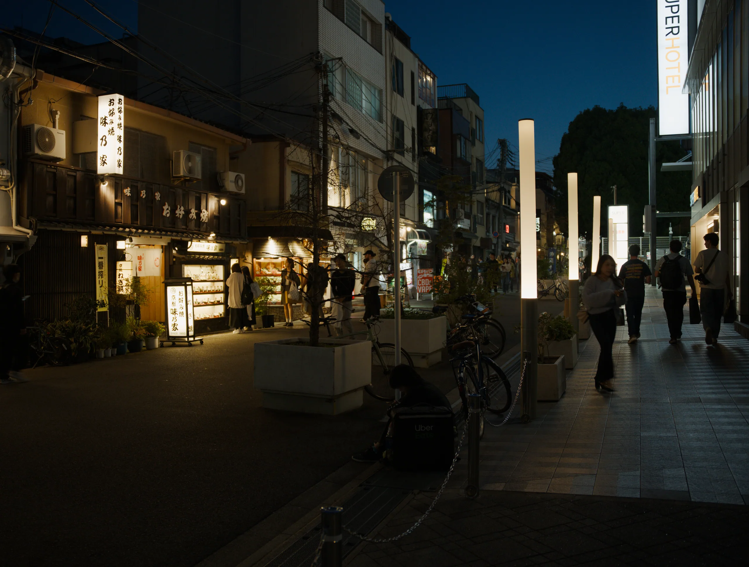 Street lined with illuminated shopfronts at dusk, a tall white column dividing the view.
