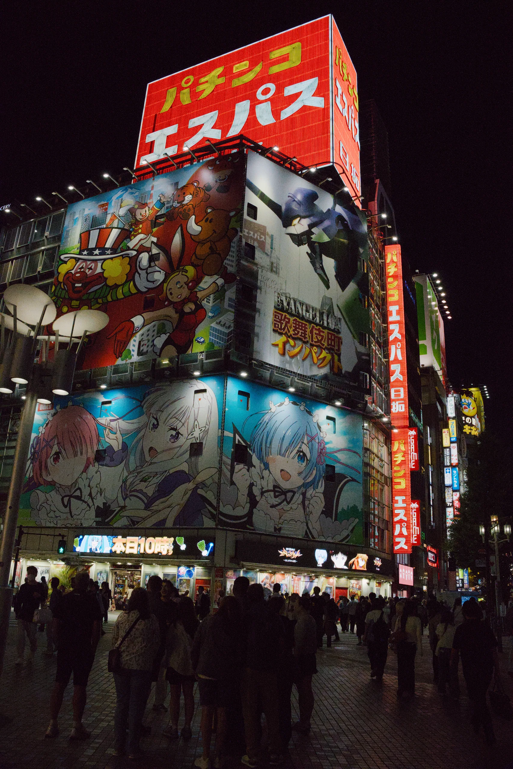 A pachinko parlour lit up at night, its facade covered floor to roof in illuminated advertising panels and a red sign in Japanese, with a crowd gathered on the street below.