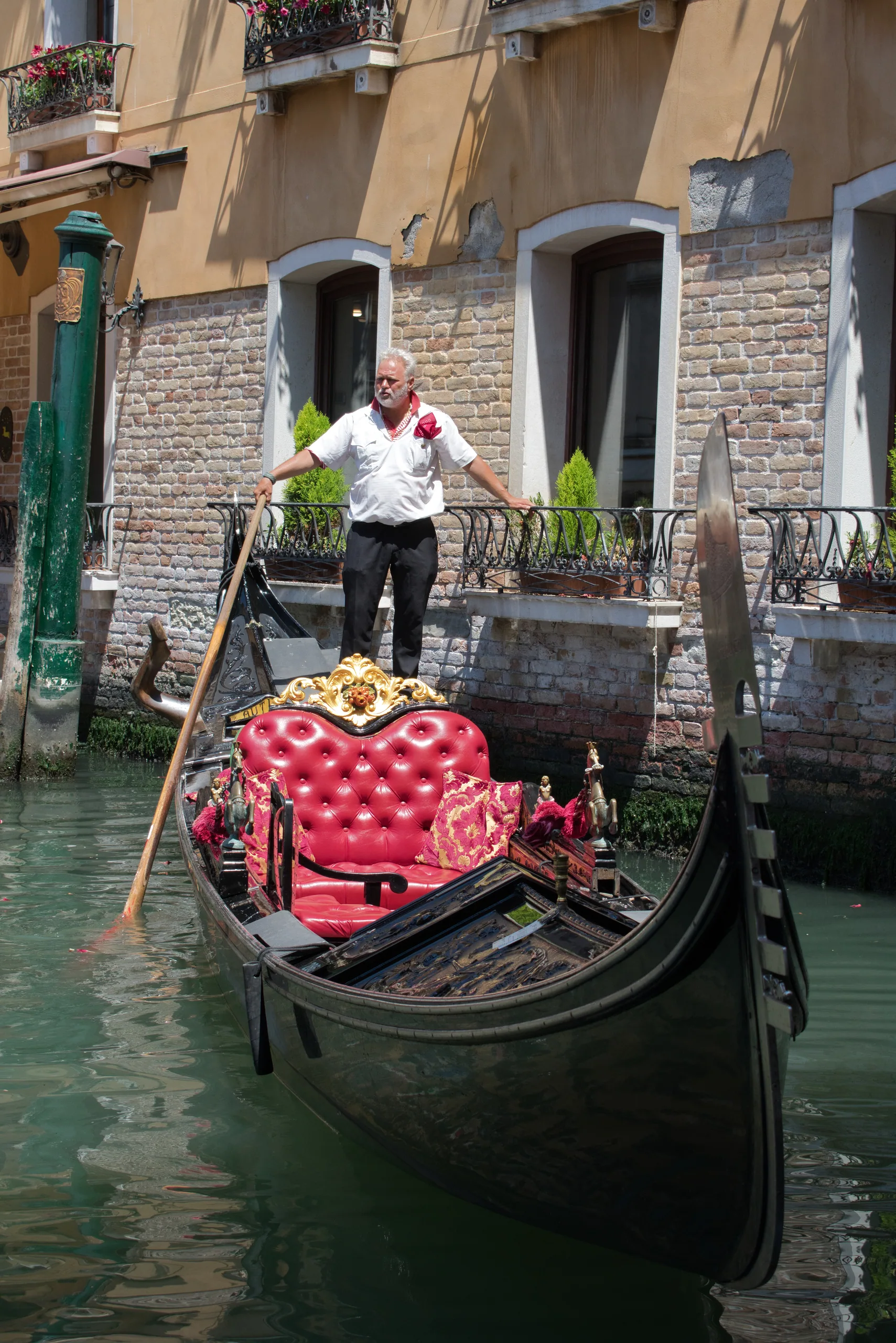 A gondolier in a white shirt standing at the stern of his gondola on a narrow canal, the boat's red velvet seats in the foreground.