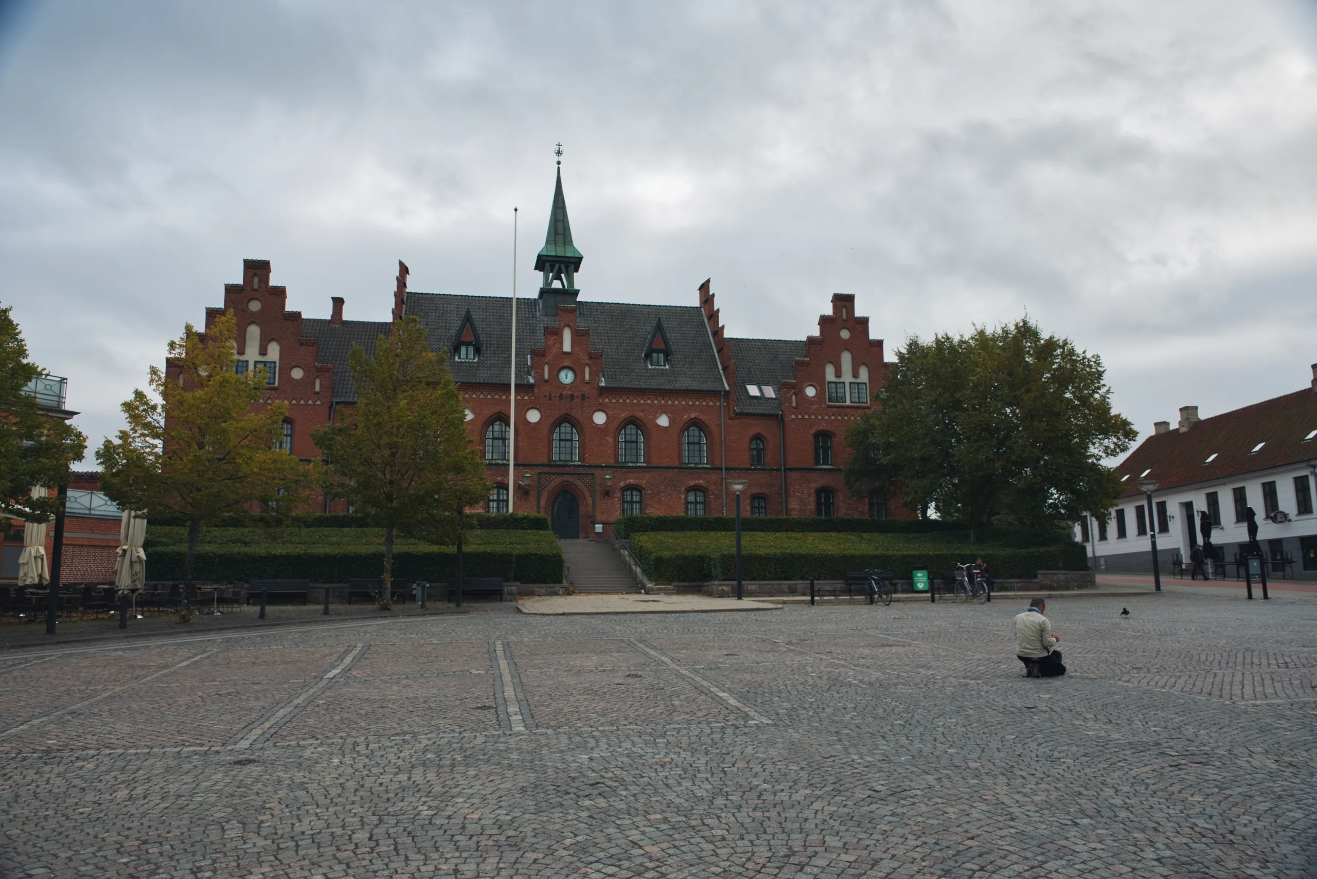 A wide cobbled square in front of a red-brick civic building with a green spire and clock, under heavy grey cloud.
