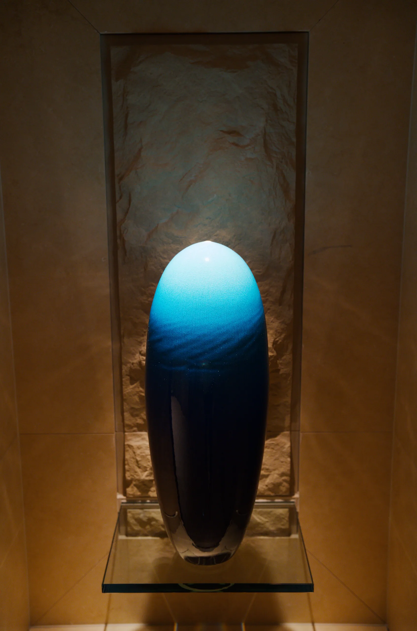 A smooth black sculptural form sits in a glass display case under blue light.