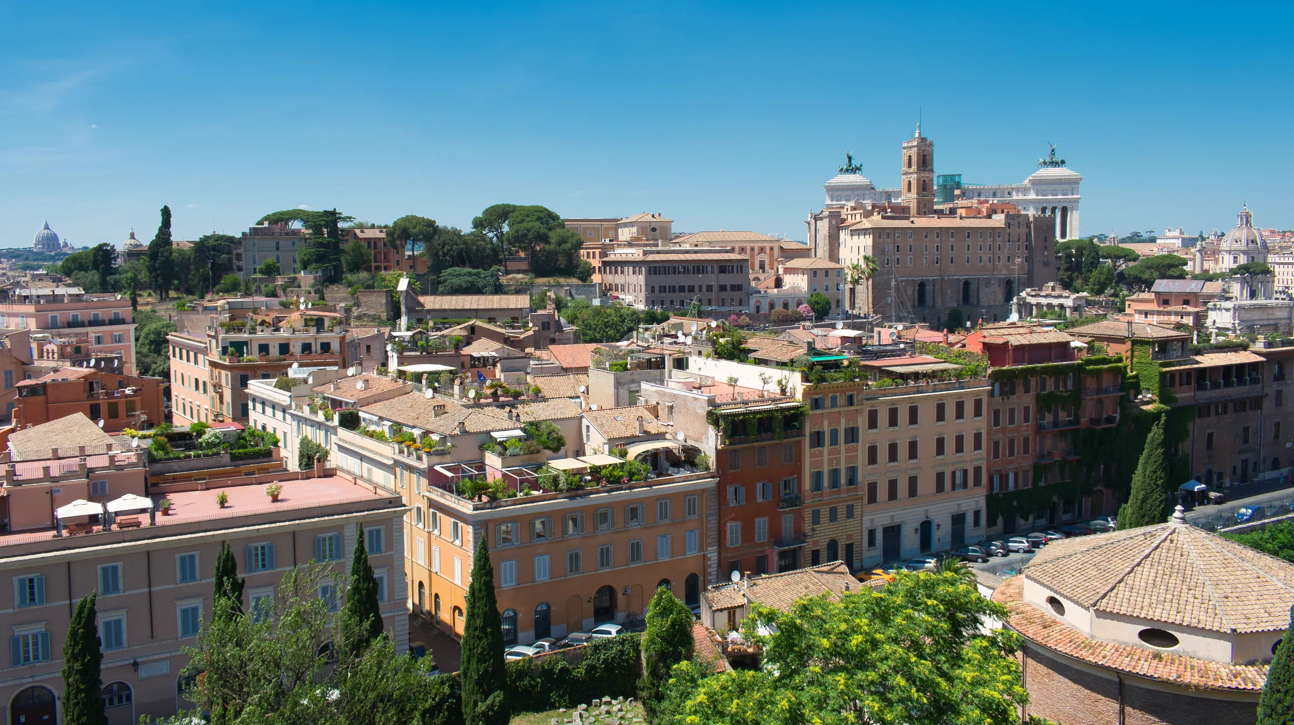 Rooftops and terraces of central Rome from above, ochre and cream buildings running back toward the Vittoriano.
