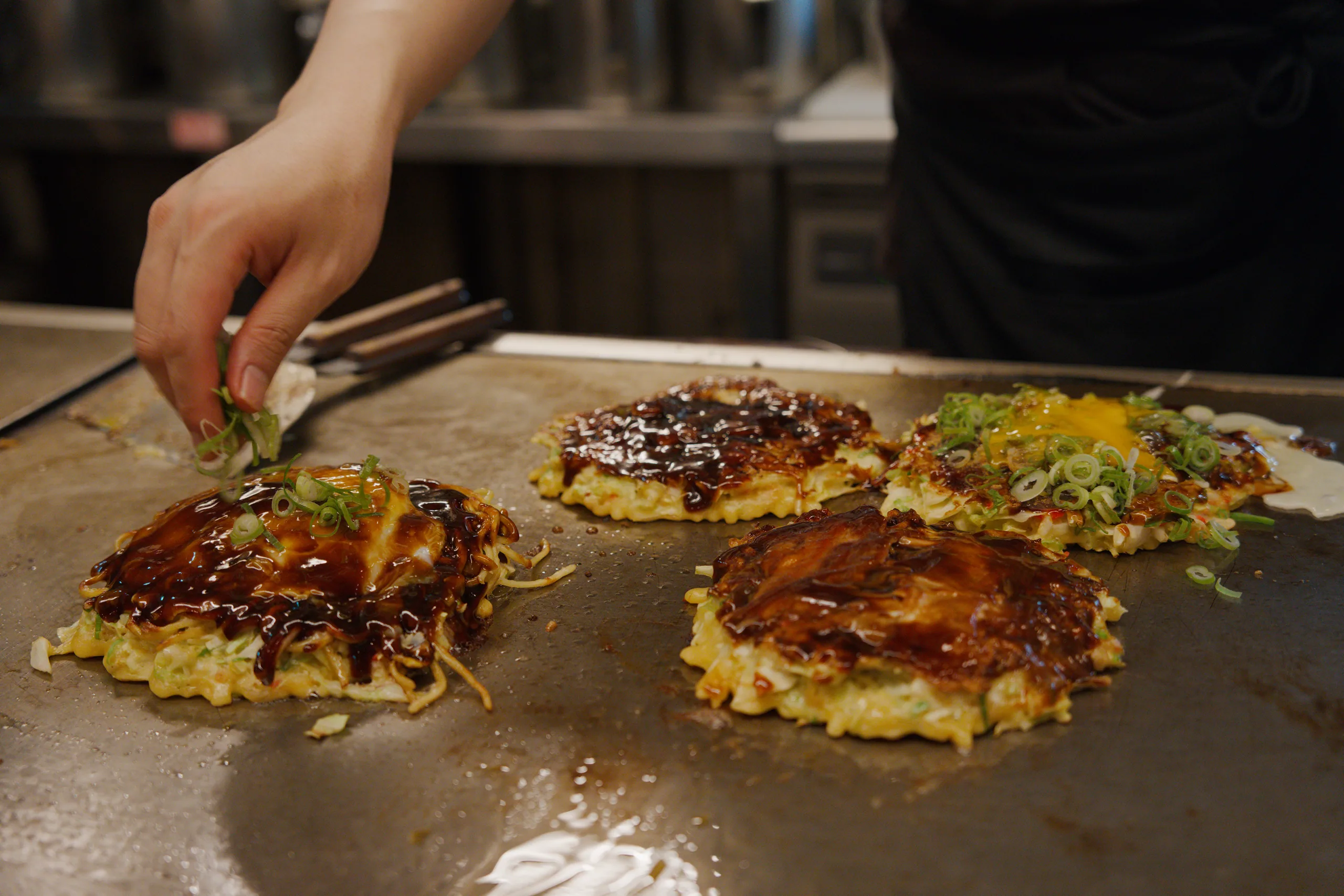 Several okonomiyaki cooking on a griddle at various stages with hands handling them.
