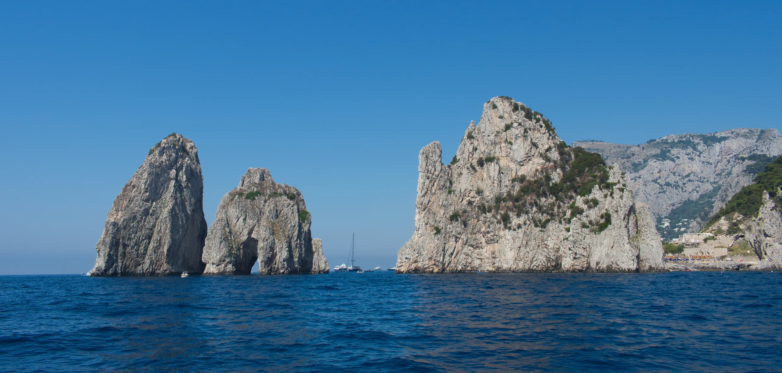 The Faraglioni sea stacks standing off Capri in deep blue water, the cliffs of the island rising behind them under a clear sky.