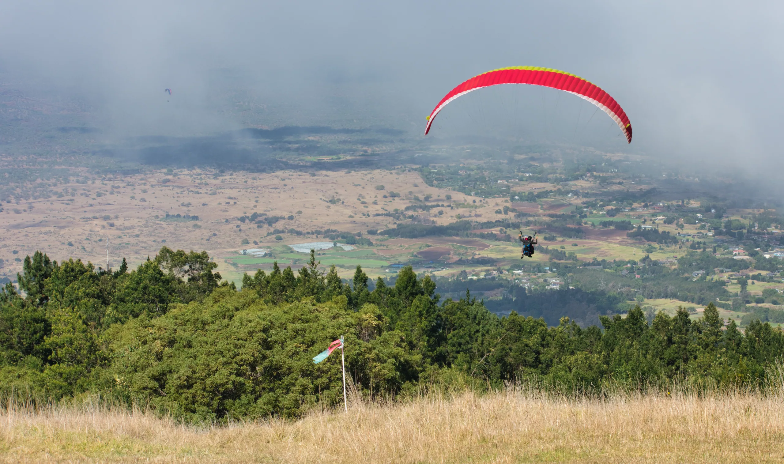 A red paraglider canopy over a patchwork of fields far below, launched from a grassy ridge in the foreground.