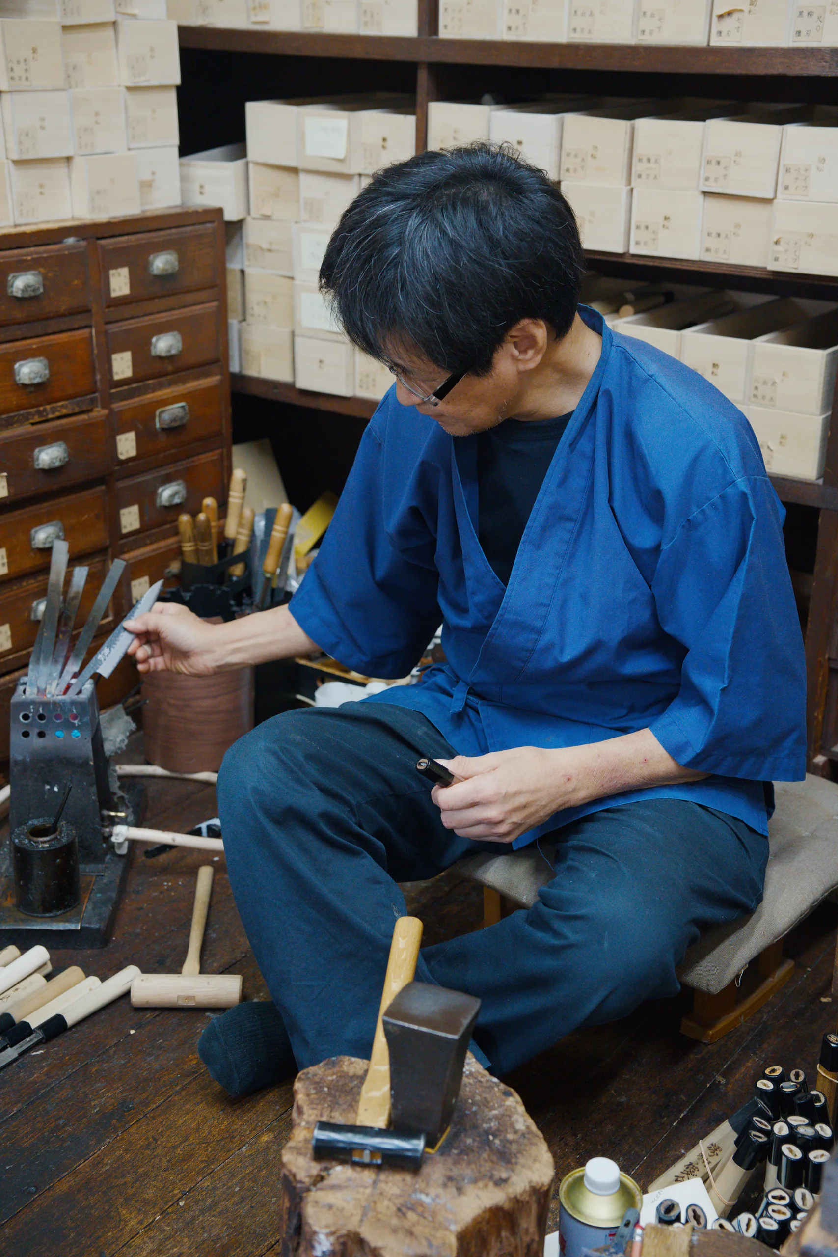 Person in blue seated and concentrating on detailed work with hand tools and materials.