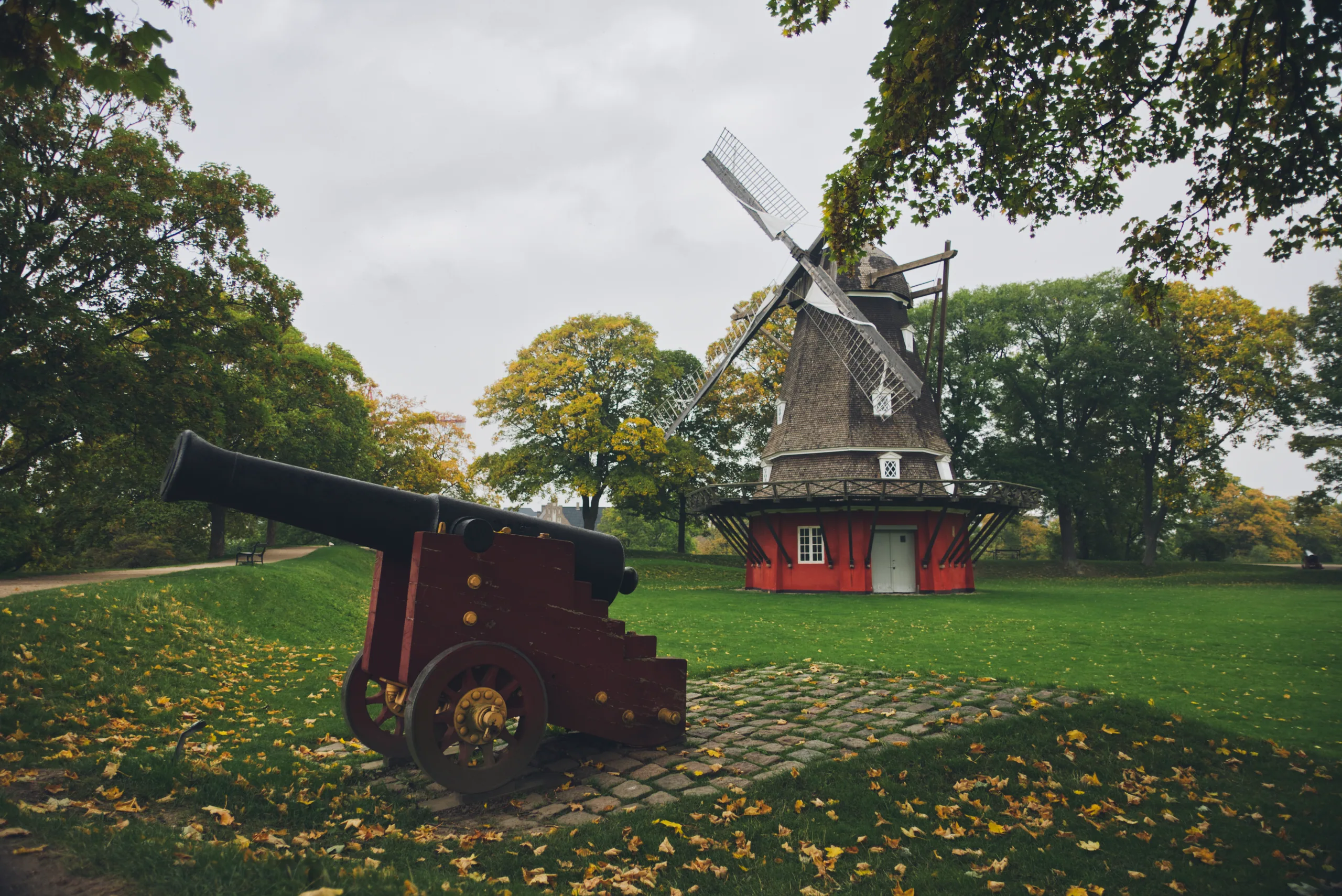 A black cannon on a red carriage on a leaf-strewn lawn, with a dark-capped windmill on a red base behind it.