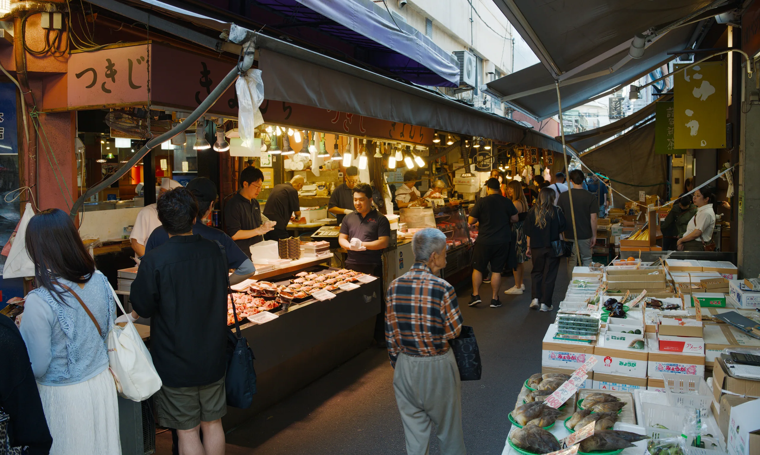 Shoppers browsing vendor stalls in an indoor market with overhead lighting and crowded aisles.
