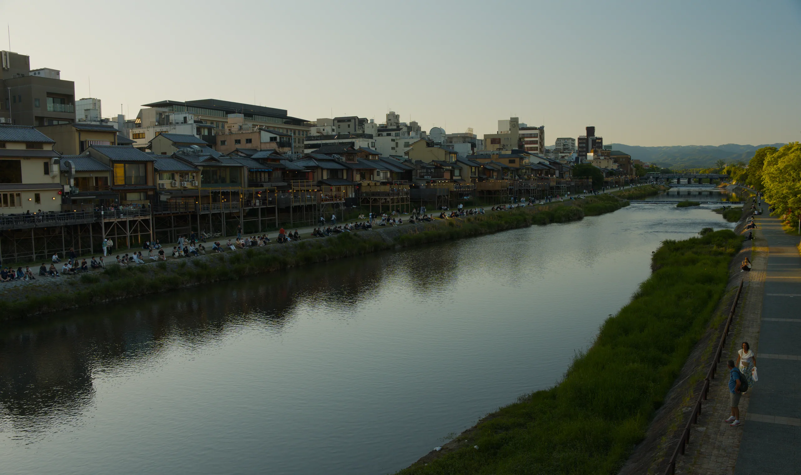Town buildings line both banks of a calm river with green hills rising in the distance.