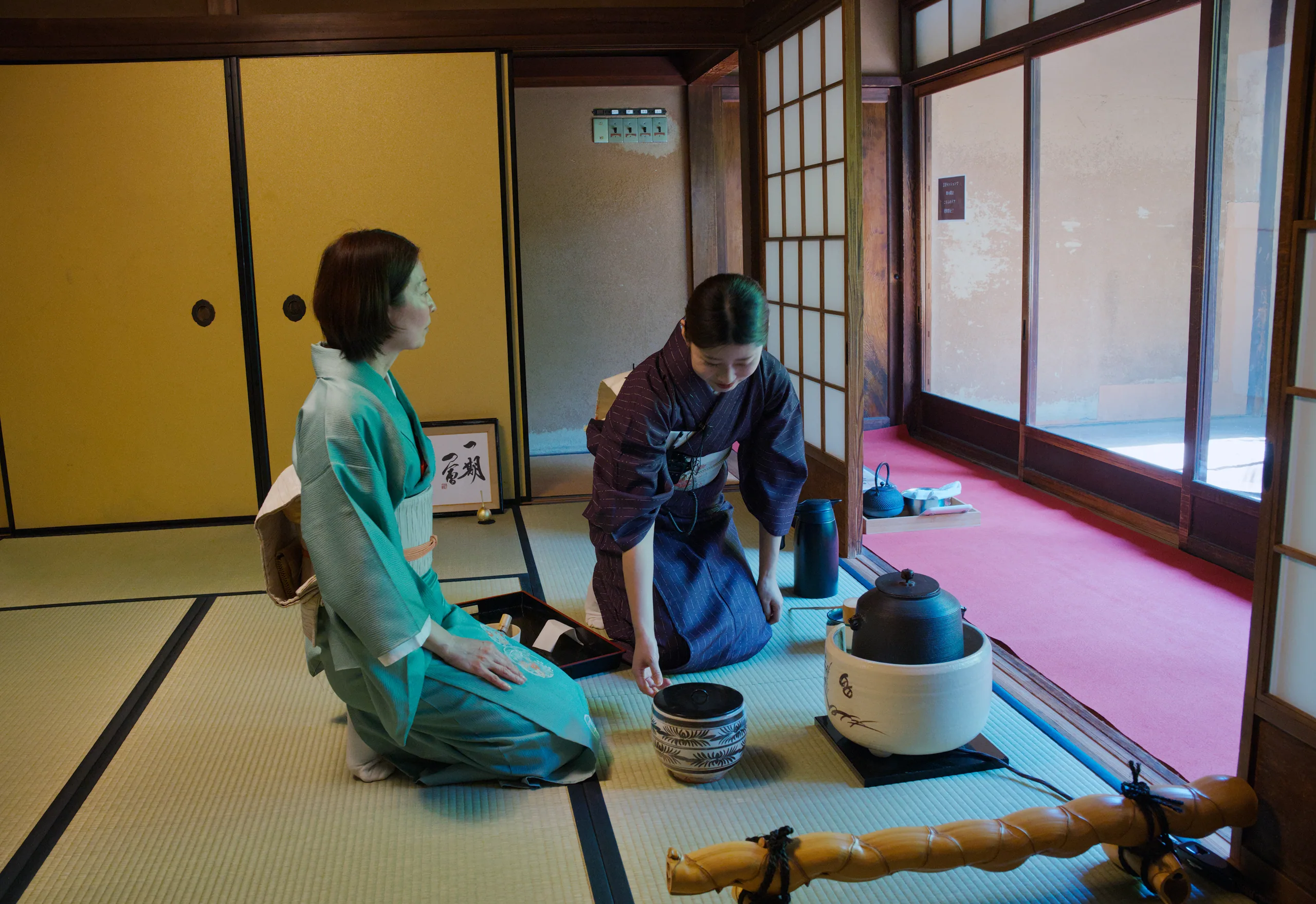 Two people in kimonos sit on tatami in a traditional Japanese room with a tea service before them.