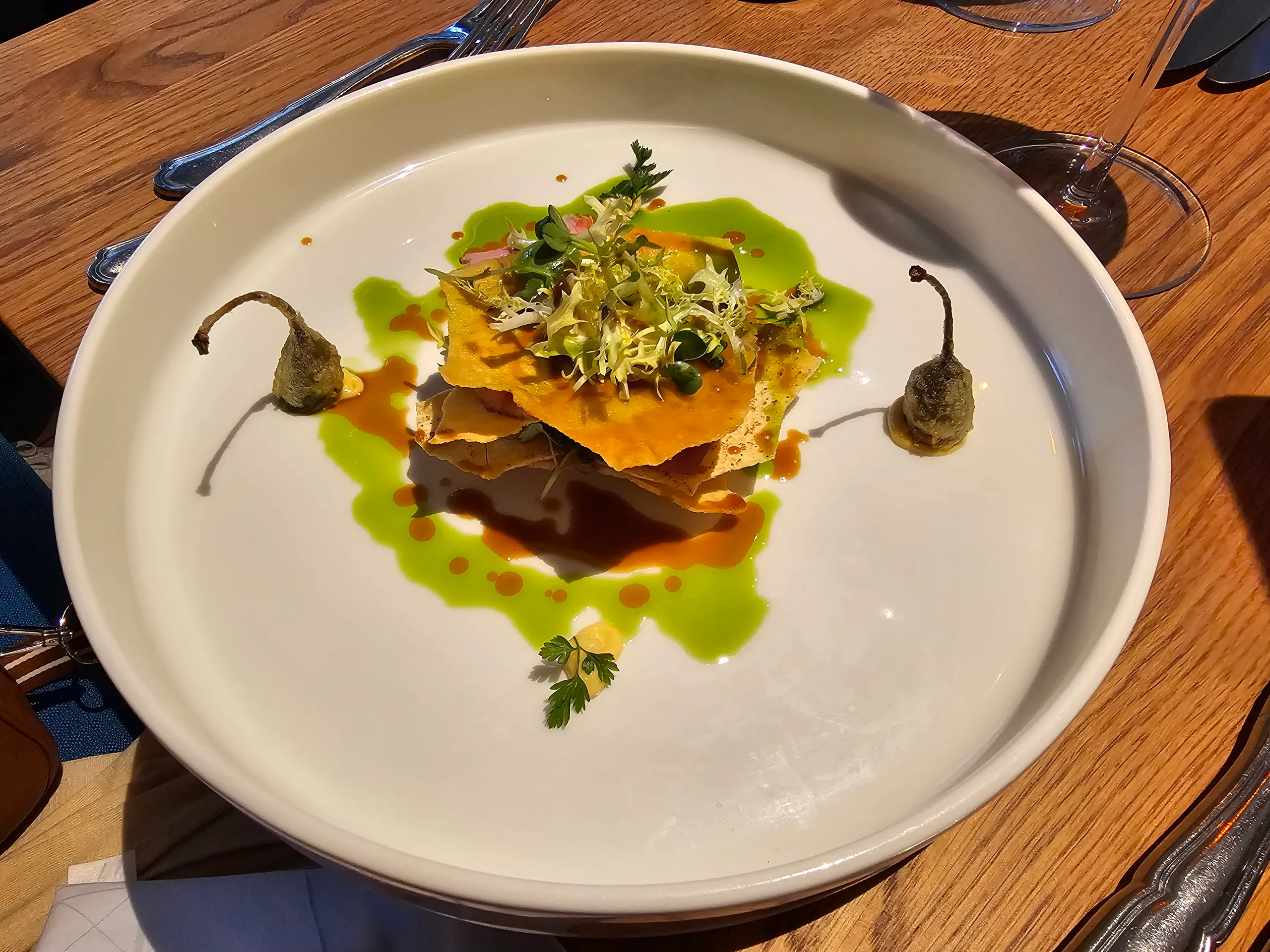 A plated dish of layered crisp pastry with herbs and capers in a pool of dark sauce and green oil.