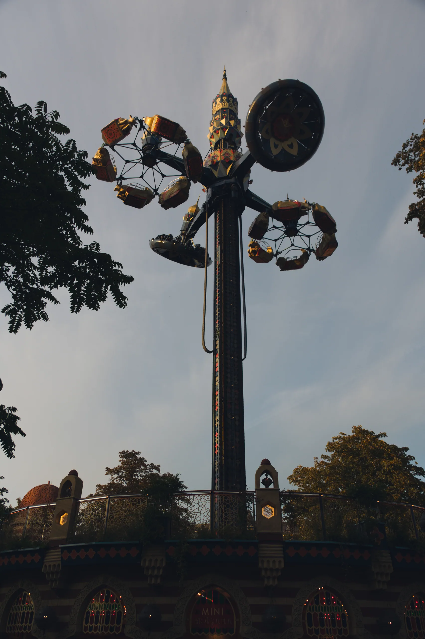 The top of the Star Flyer tower at Tivoli against a pale evening sky, framed by dark leaves.