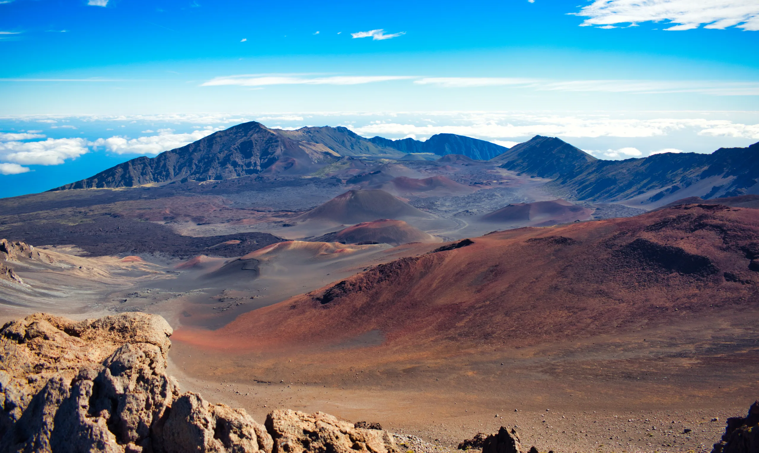 The red and ochre cinder cones of Haleakalā crater seen from the rim, cloud banked along the far edge.