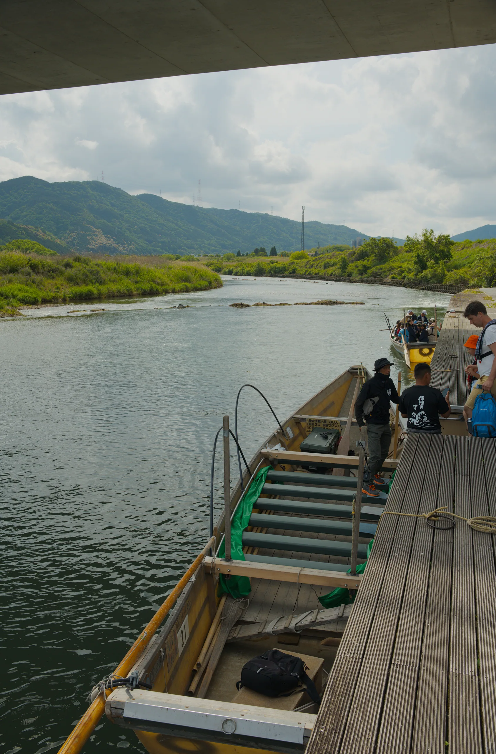 Passengers board a wooden boat at a riverside dock under mountains.