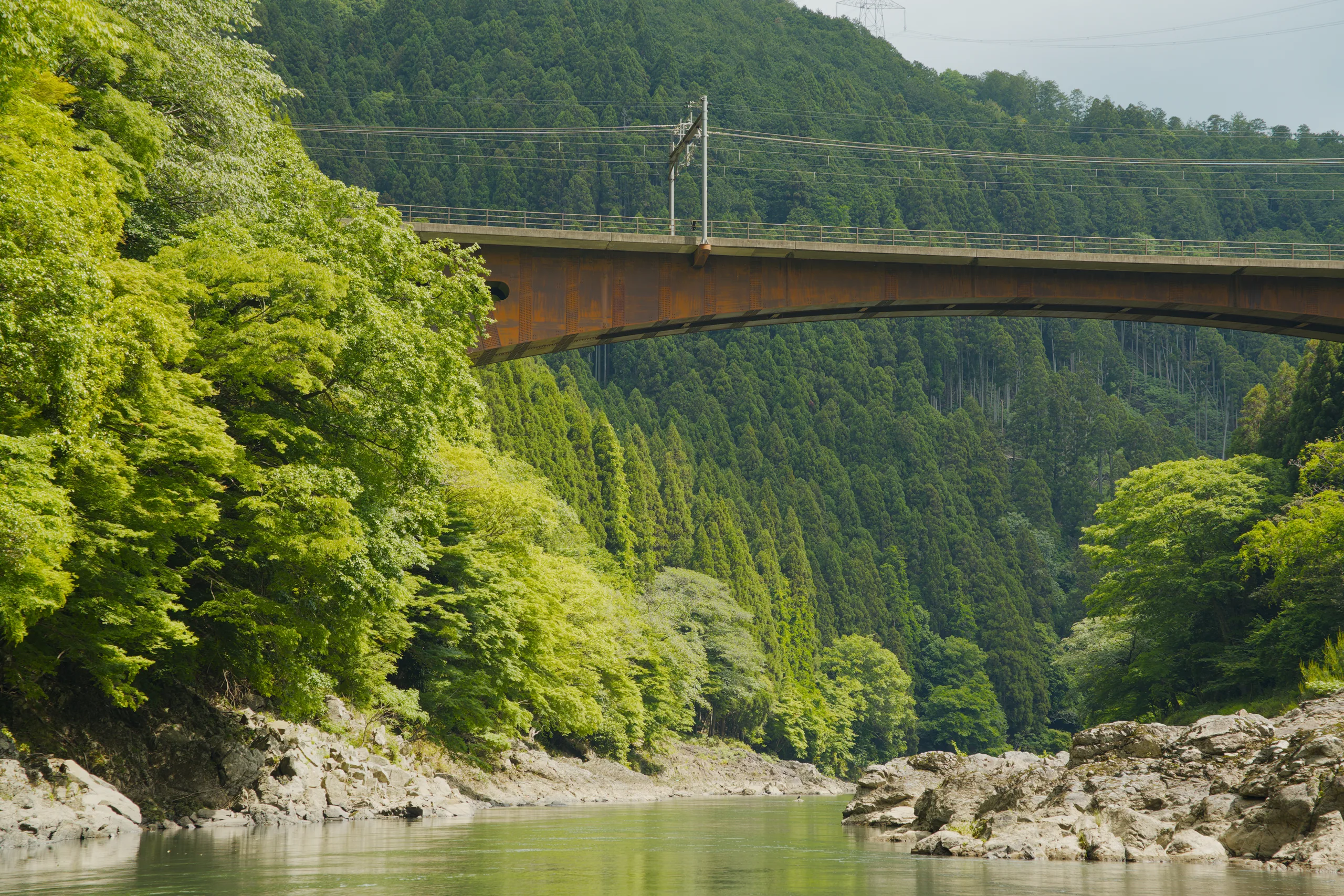 A river flows between forested slopes beneath an arching metal footbridge.