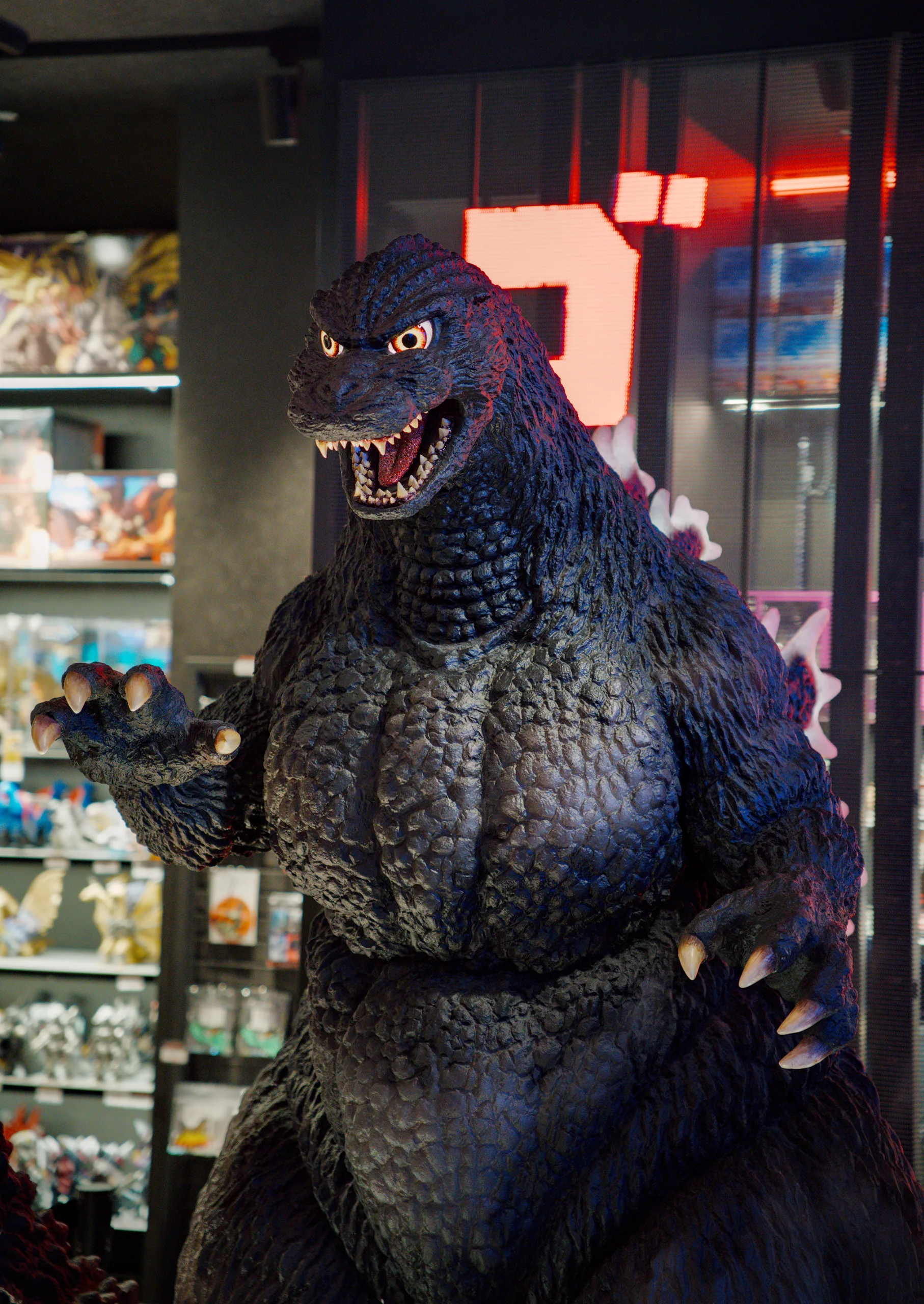 Godzilla statue with open mouth and raised arm in a glass display case with neon signage behind.