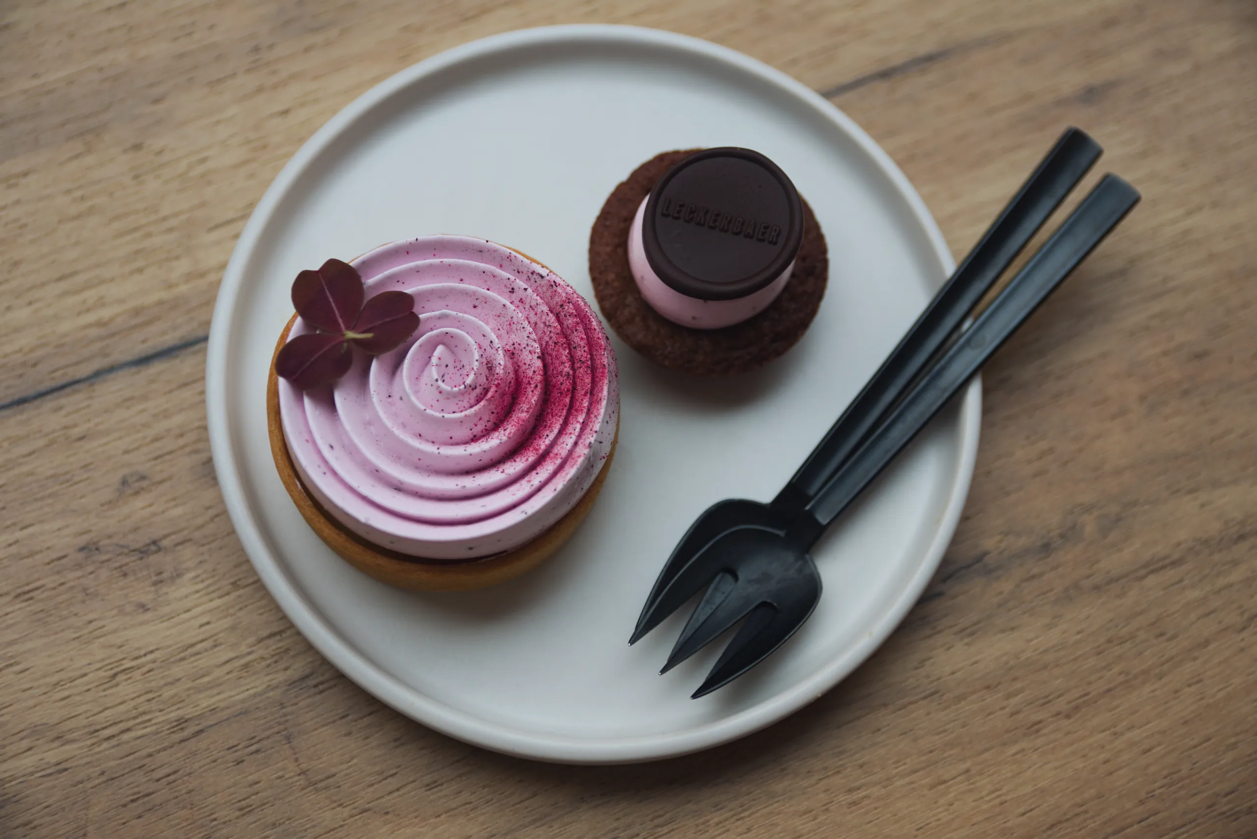 Two small cakes on a white plate with a black fork — a pink meringue-topped tart and a chocolate-glazed one — on a pale wooden table.