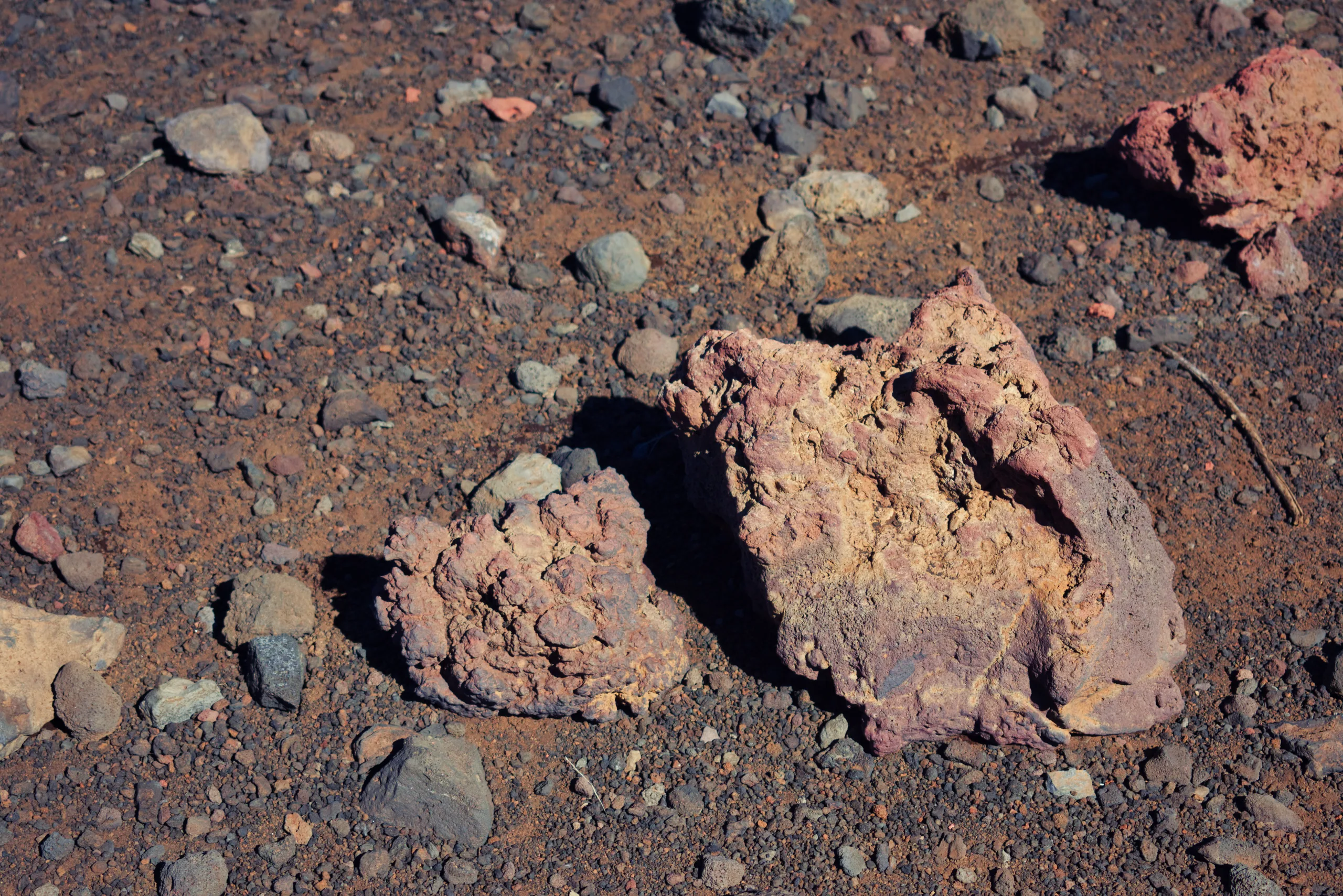 Rust-red volcanic rock and cinder filling the frame, one larger block casting a hard shadow.