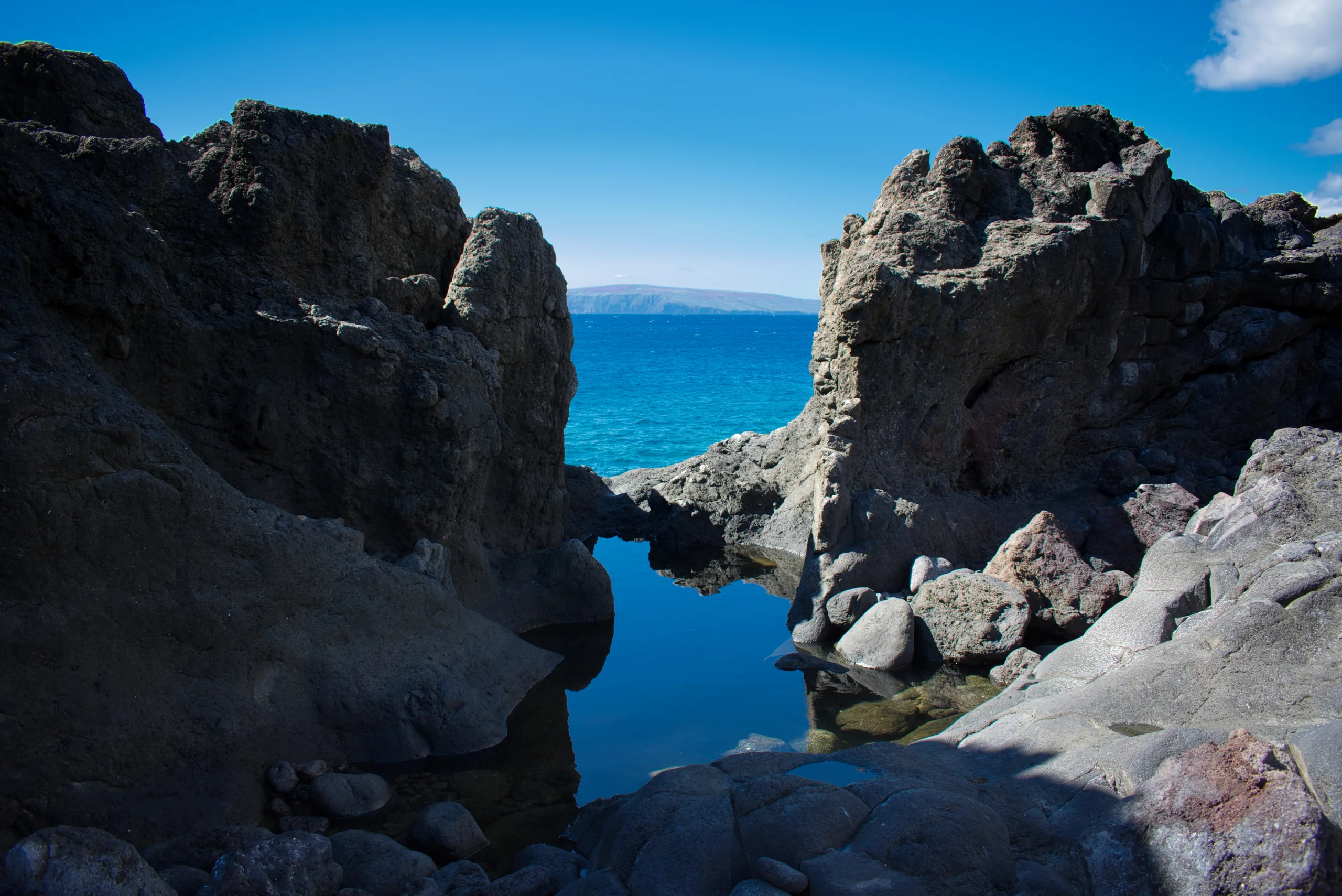 A still tidal pool held between black lava outcrops, the open ocean and a distant island visible through the gap.