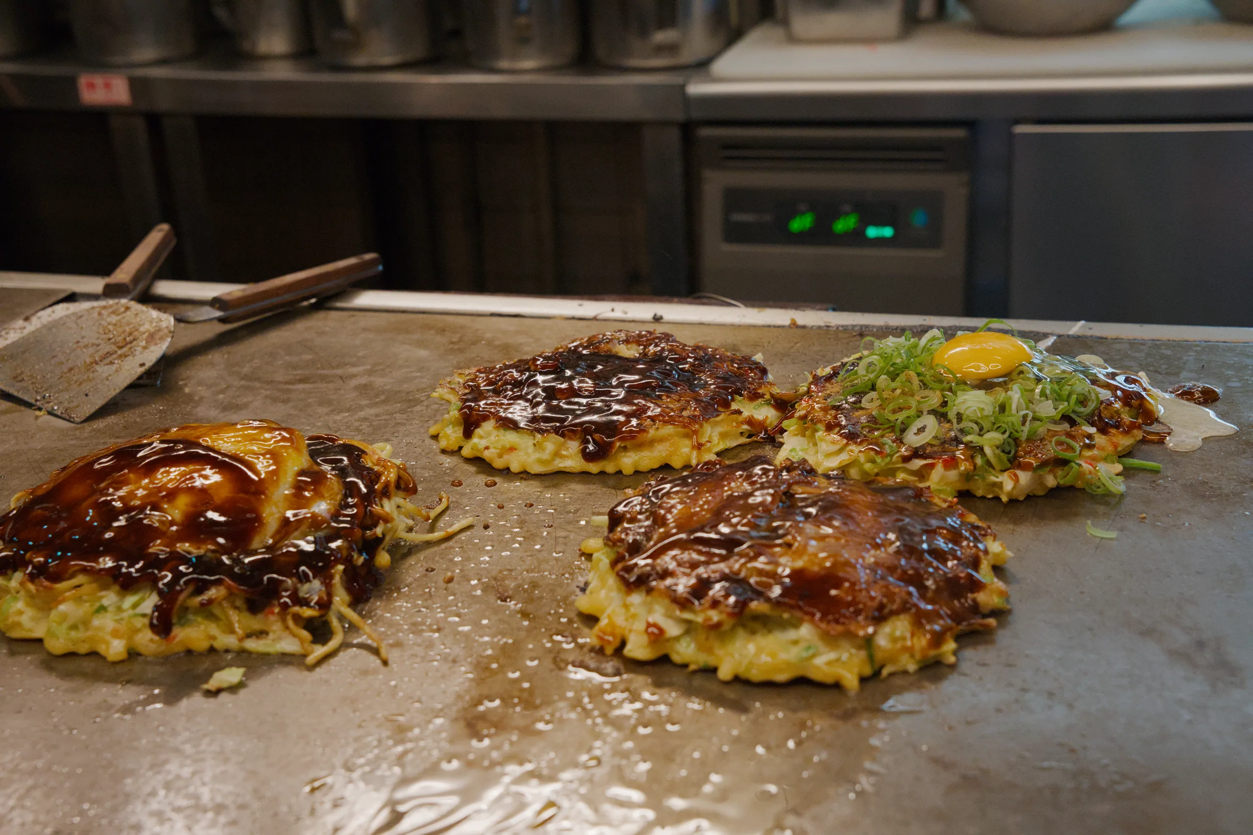 Hands with metal spatulas working okonomiyaki on a cooking griddle.