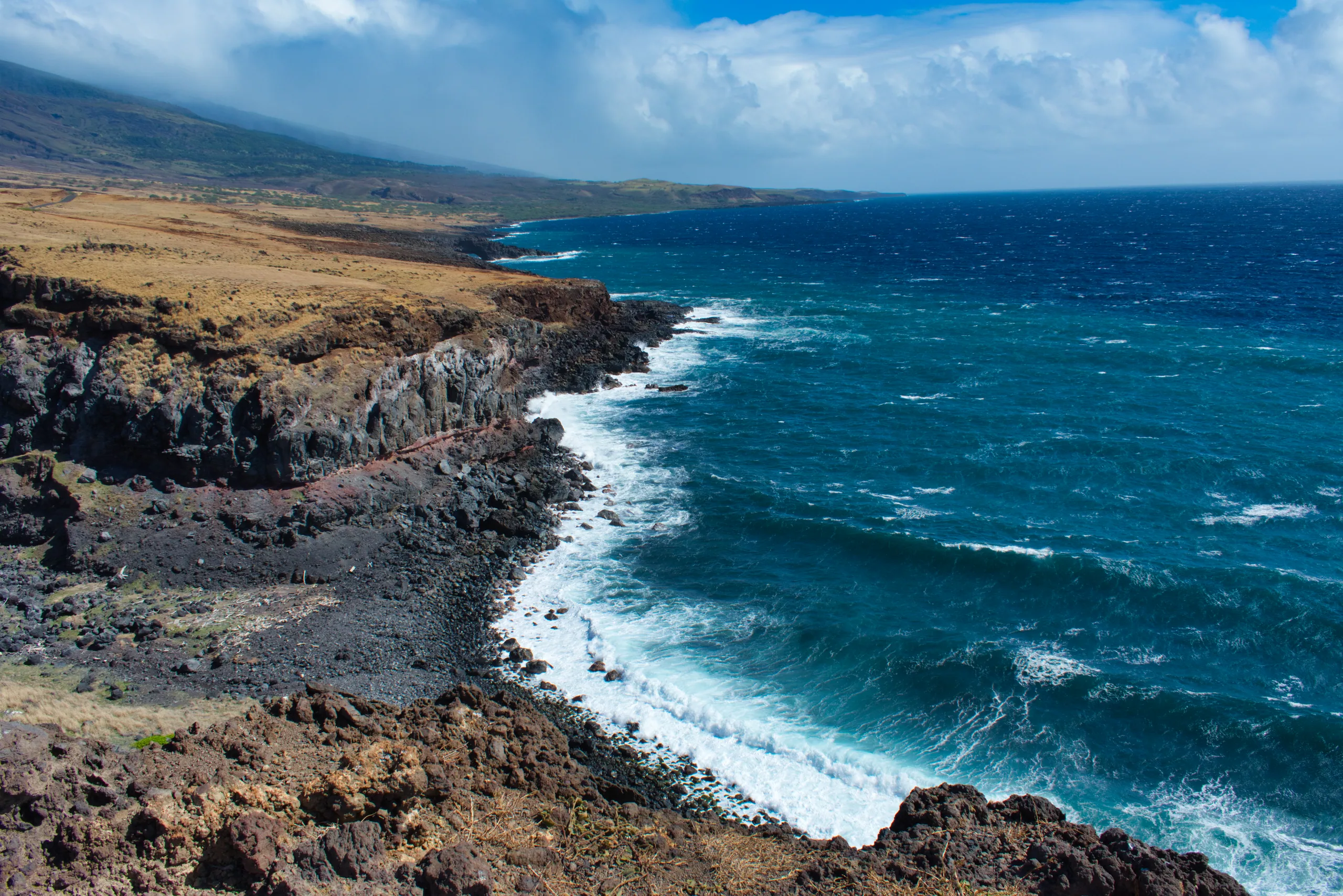 Surf breaking white along a rugged lava coastline, deep blue ocean beyond and dry scrubland in the foreground.