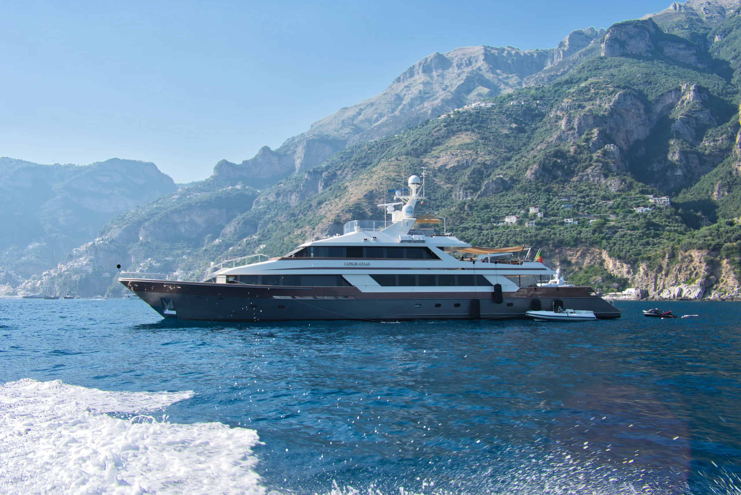 A large motor yacht running along the Amalfi coast, terraced mountains rising steeply behind it.