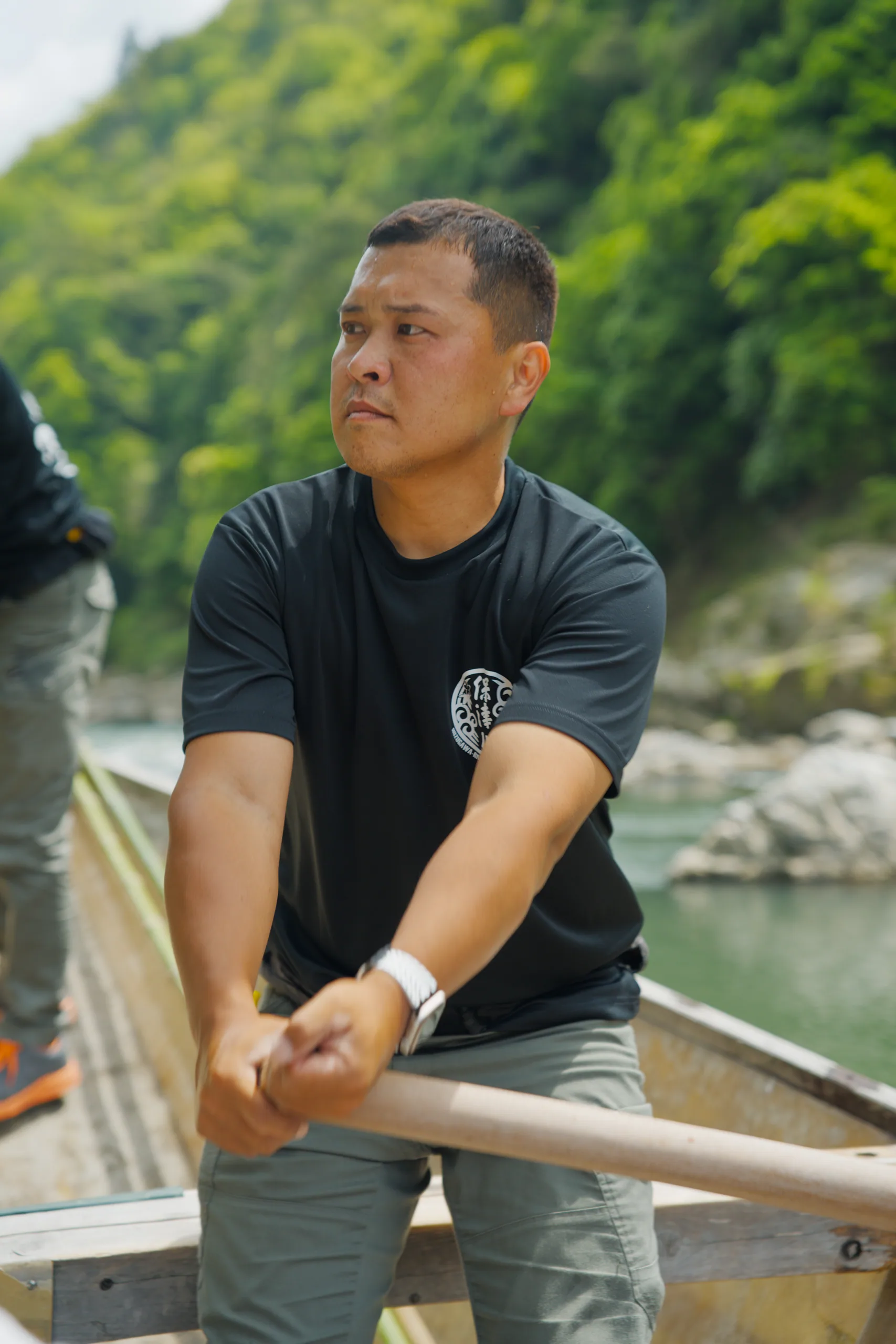 A man seated in a boat grips a paddle, with forested banks behind him.