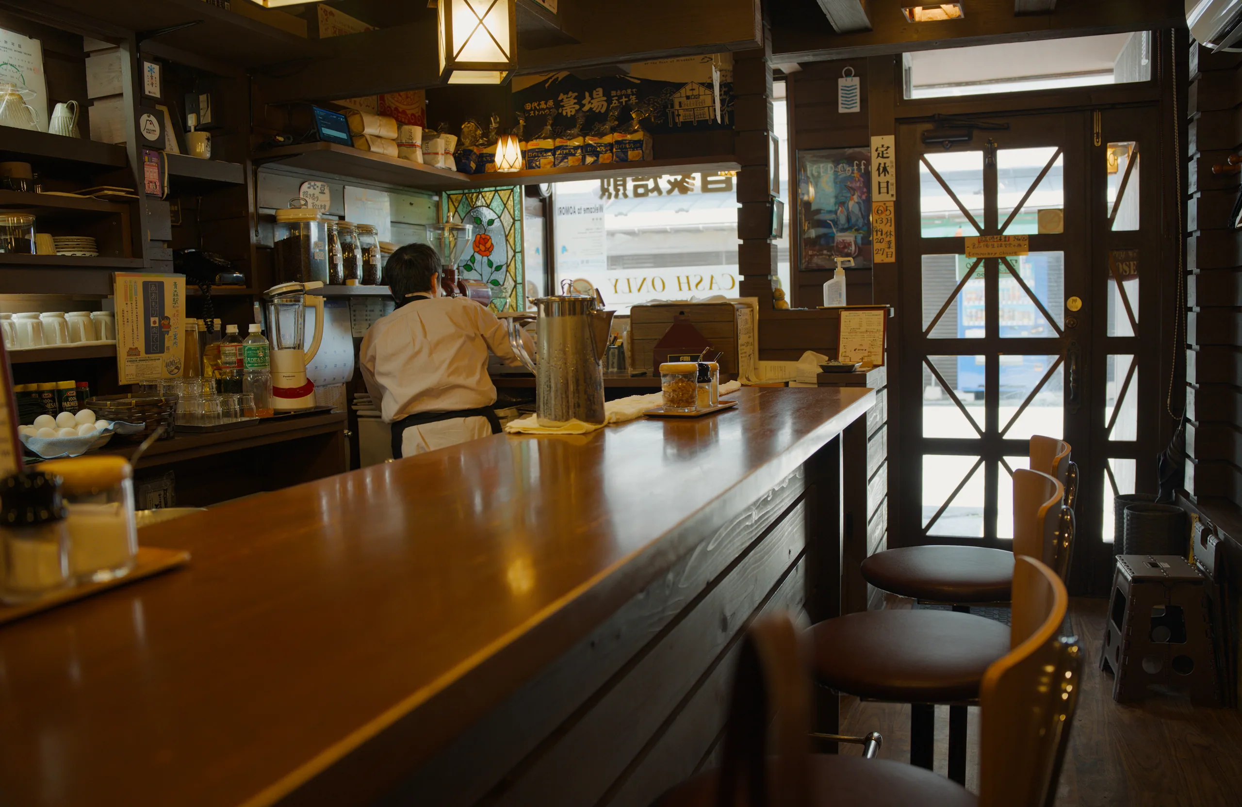 A server stands behind a wooden counter in a warmly lit restaurant with cross-hatched window frames.