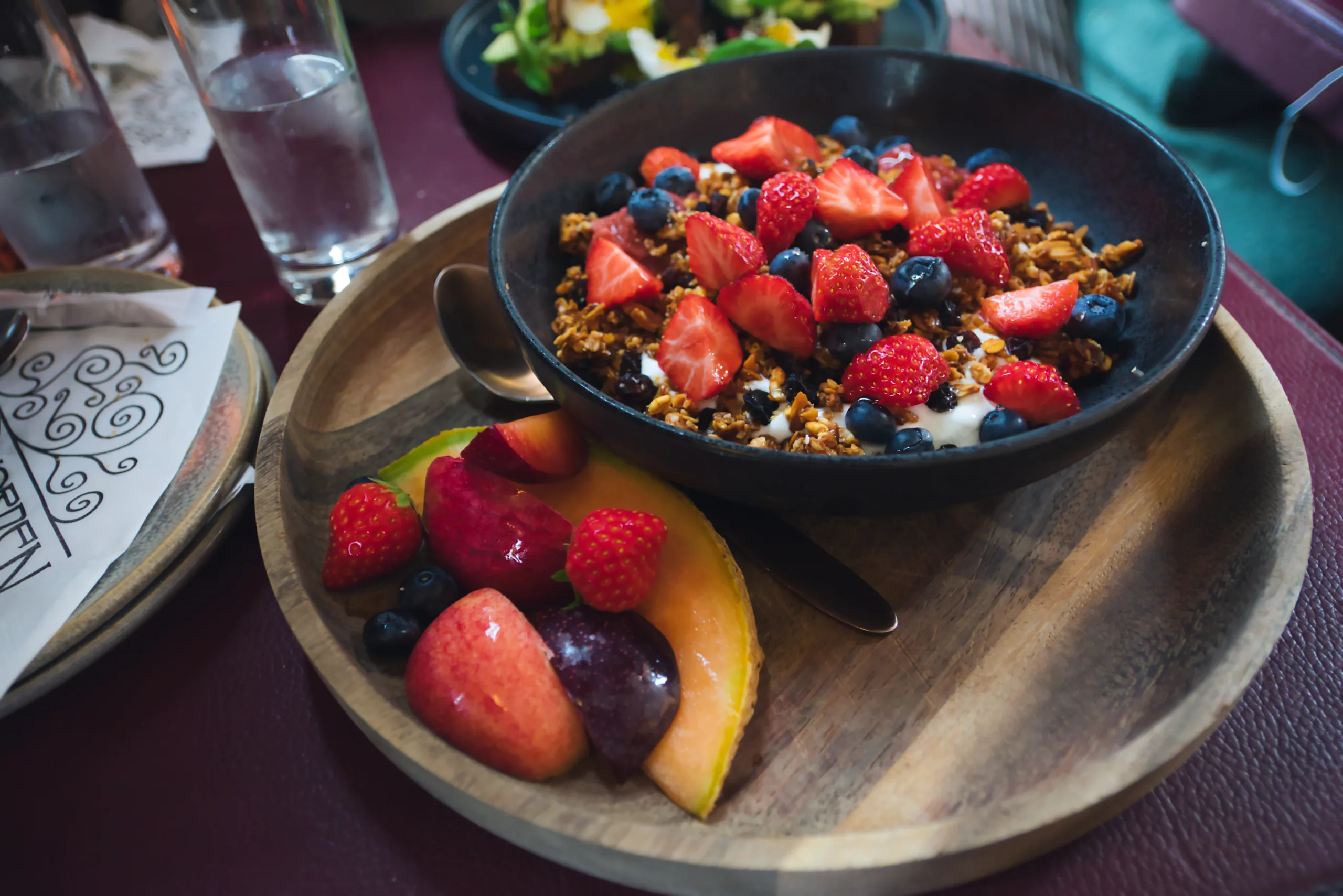 A breakfast of granola, strawberries and blueberries in a dark bowl, with a plate of cut melon and berries beside it.