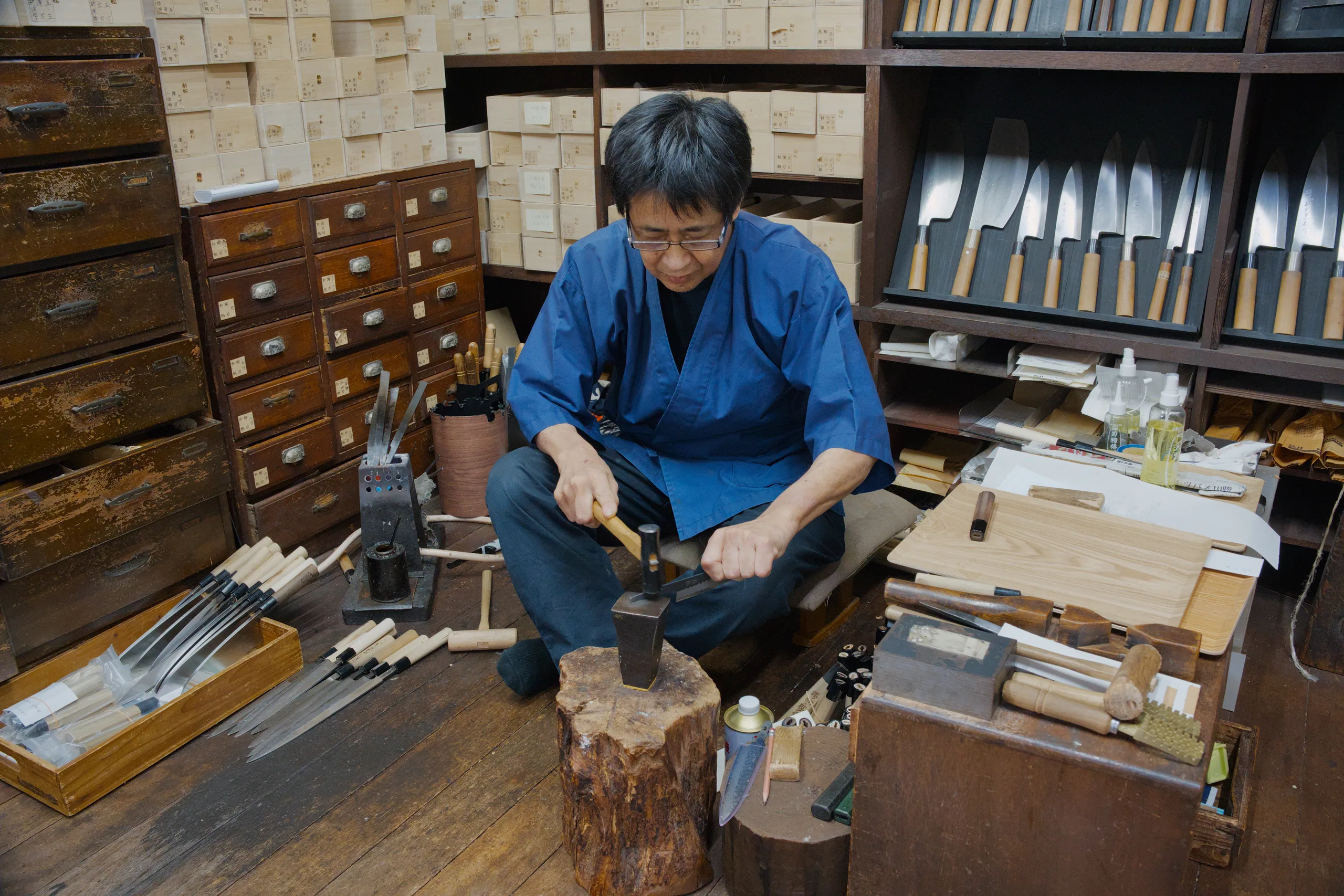 Person in blue crouching at a work station with hand tools and wooden materials, demonstrating a traditional craft.