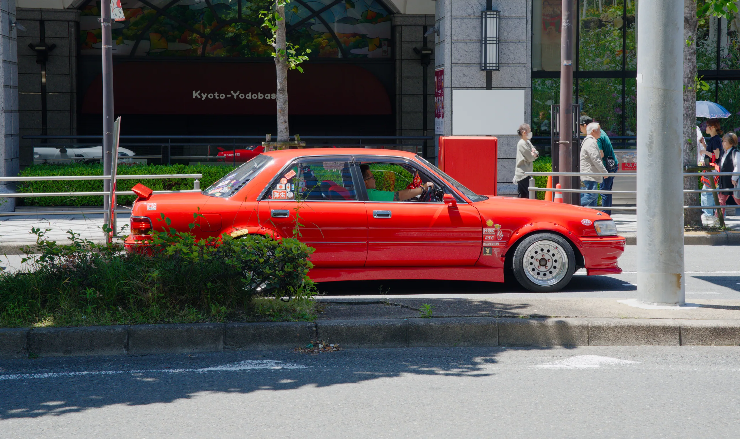 A red car parked on a street in front of an ivy-covered building, with a pedestrian nearby.