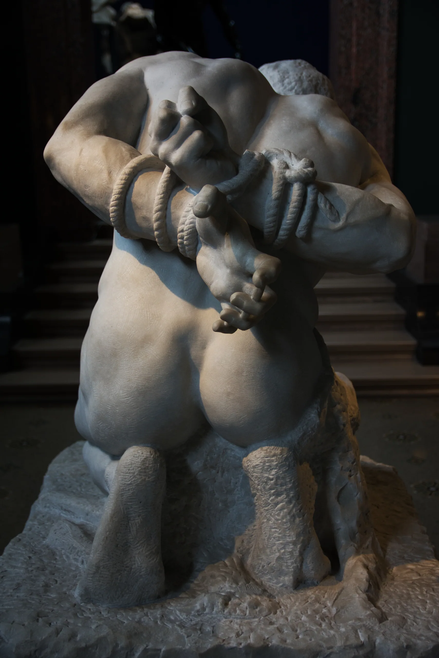 A muscular marble figure crouched on a rock with his arms bound in rope, seen from behind in a dim gallery.