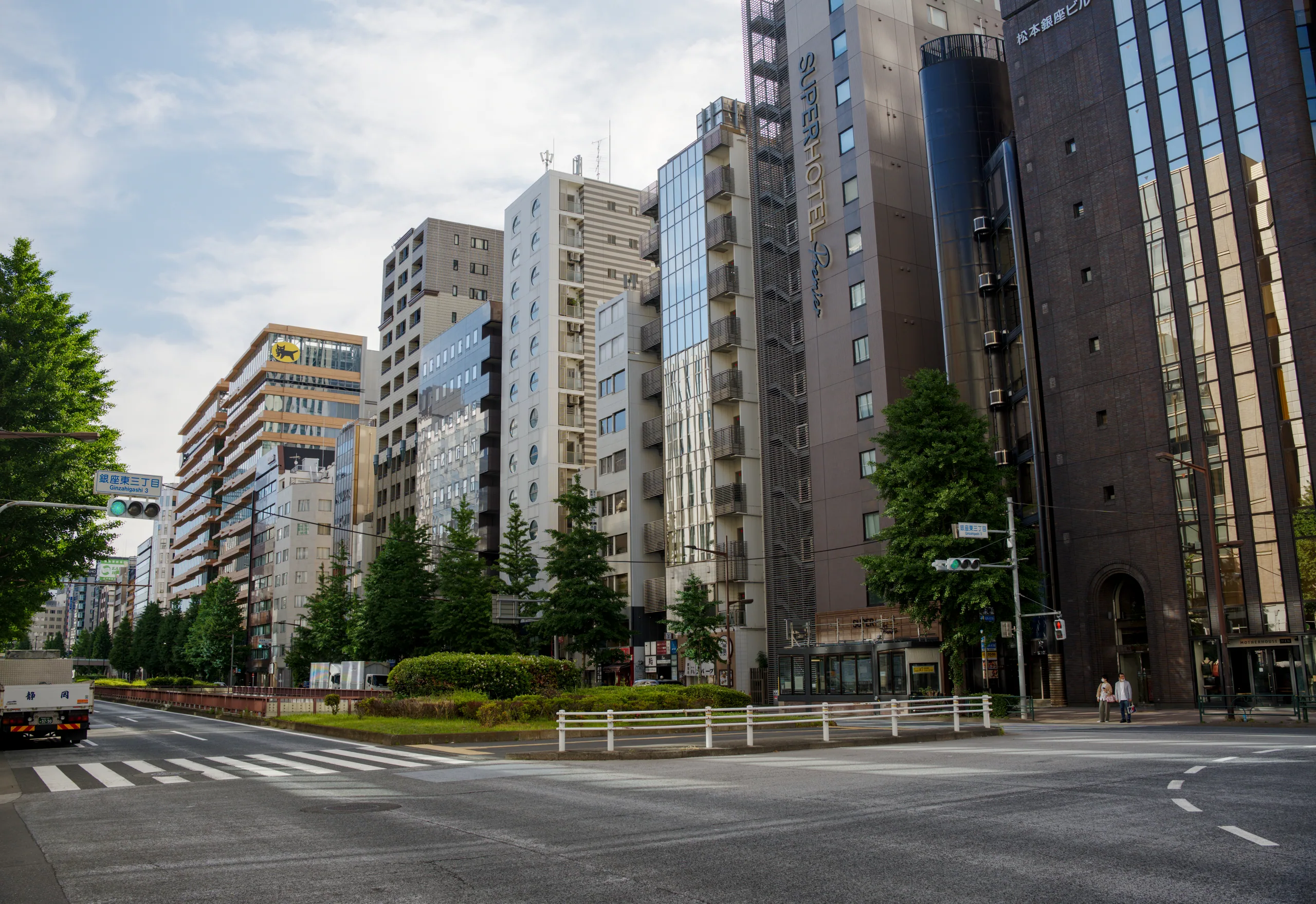 Tall office and residential towers flank a zebra crossing on a street lined with green trees.