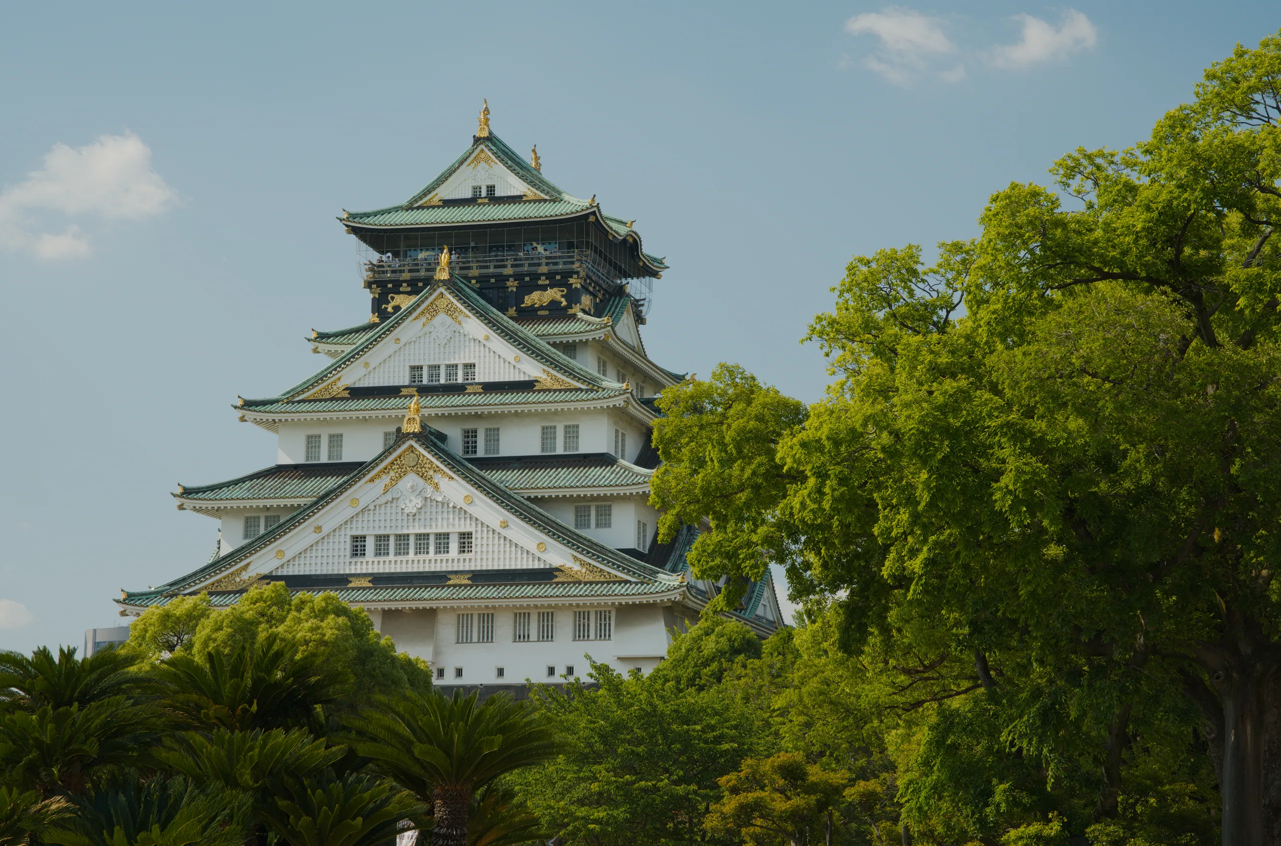 Japanese castle with stacked pagoda-like tiers and golden roof ornaments, surrounded by green foliage.