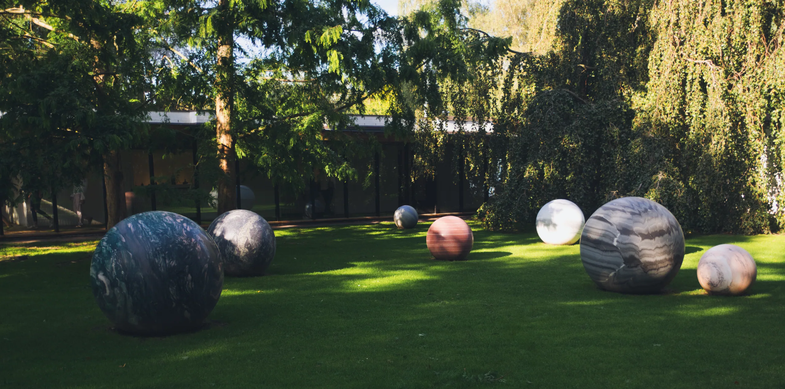 Large marbled stone spheres set out across a sunlit lawn in a sculpture garden, trees and a low gallery building behind.