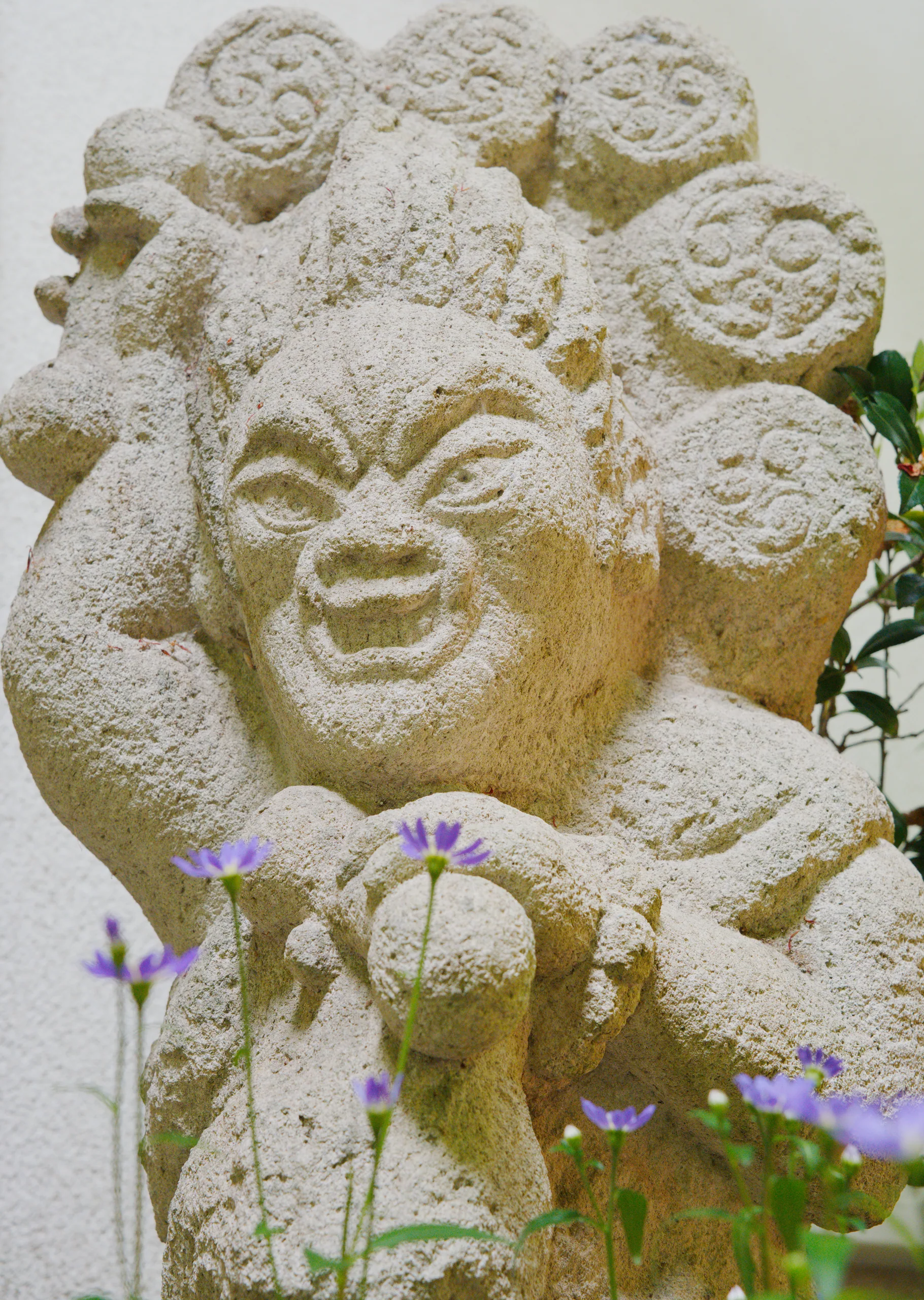 Stone sculpture with multiple carved faces and blue flowers blooming at its base.