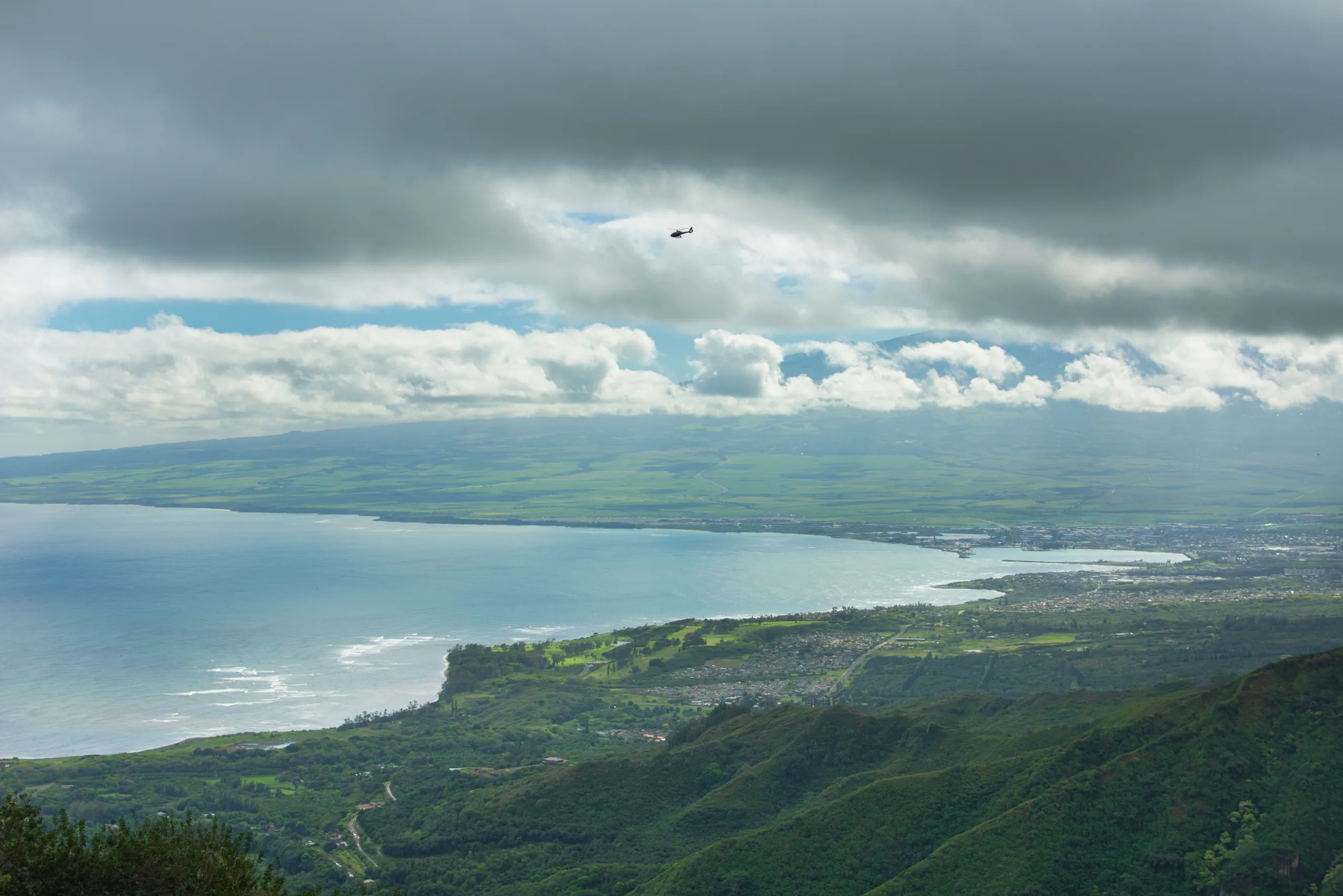 A high view across a green isthmus to the sea on both sides, under a heavy grey cloud layer.